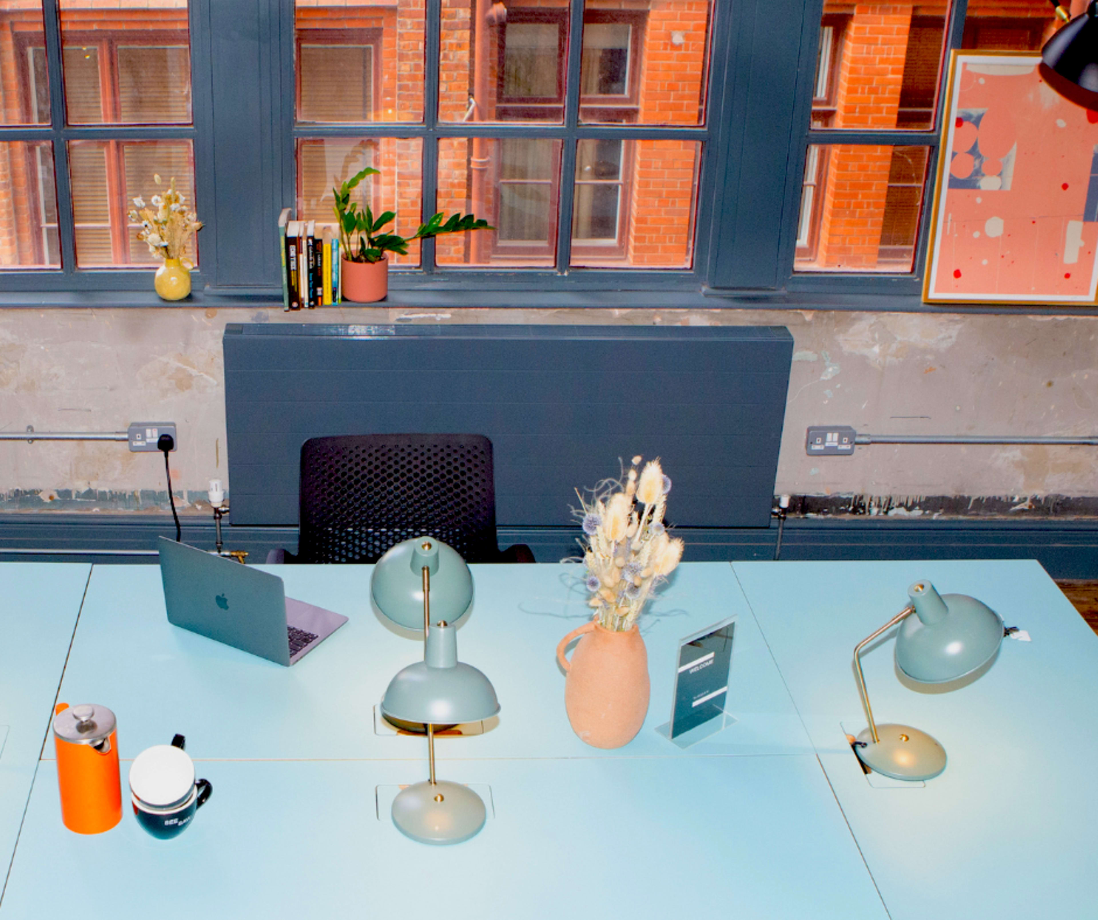 A modern office desk with a laptop, two desk lamps, a vase with dried flowers, and a coffee cup, all set against a backdrop of large windows revealing a brick building.
