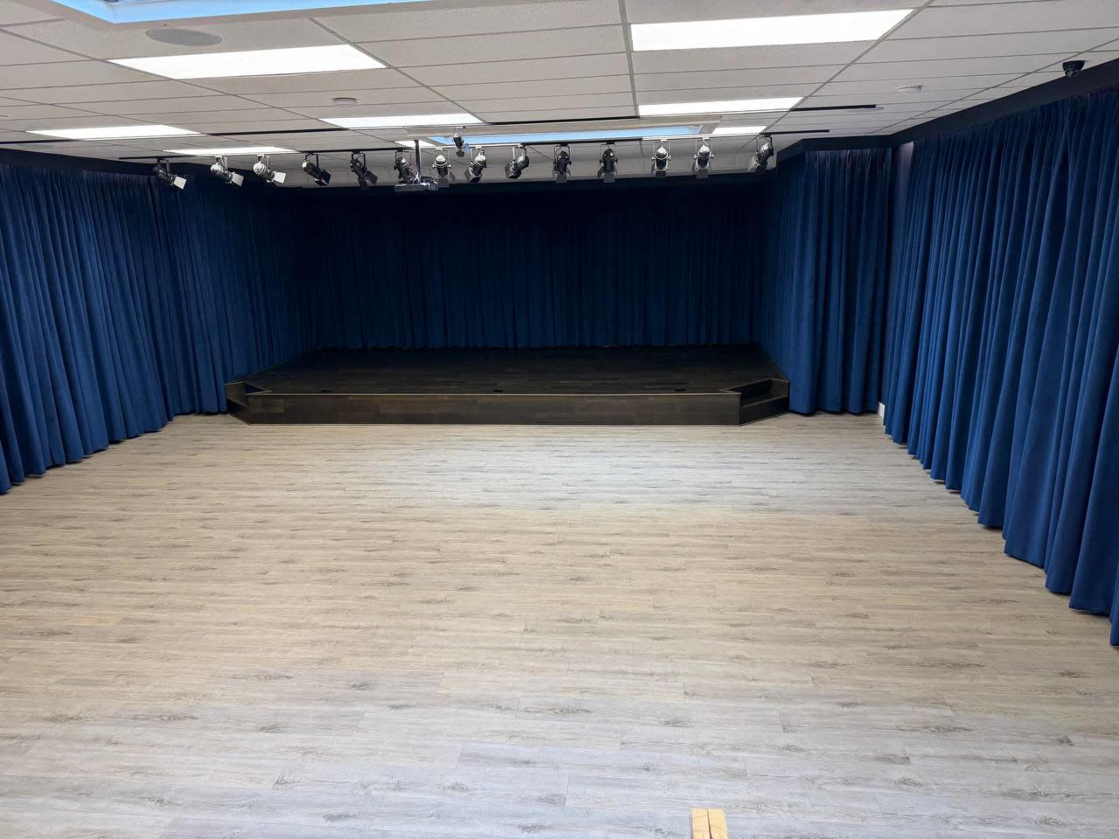 The image shows a blank performance space with a wooden stage at the back, surrounded by blue curtains and hardwood flooring.
