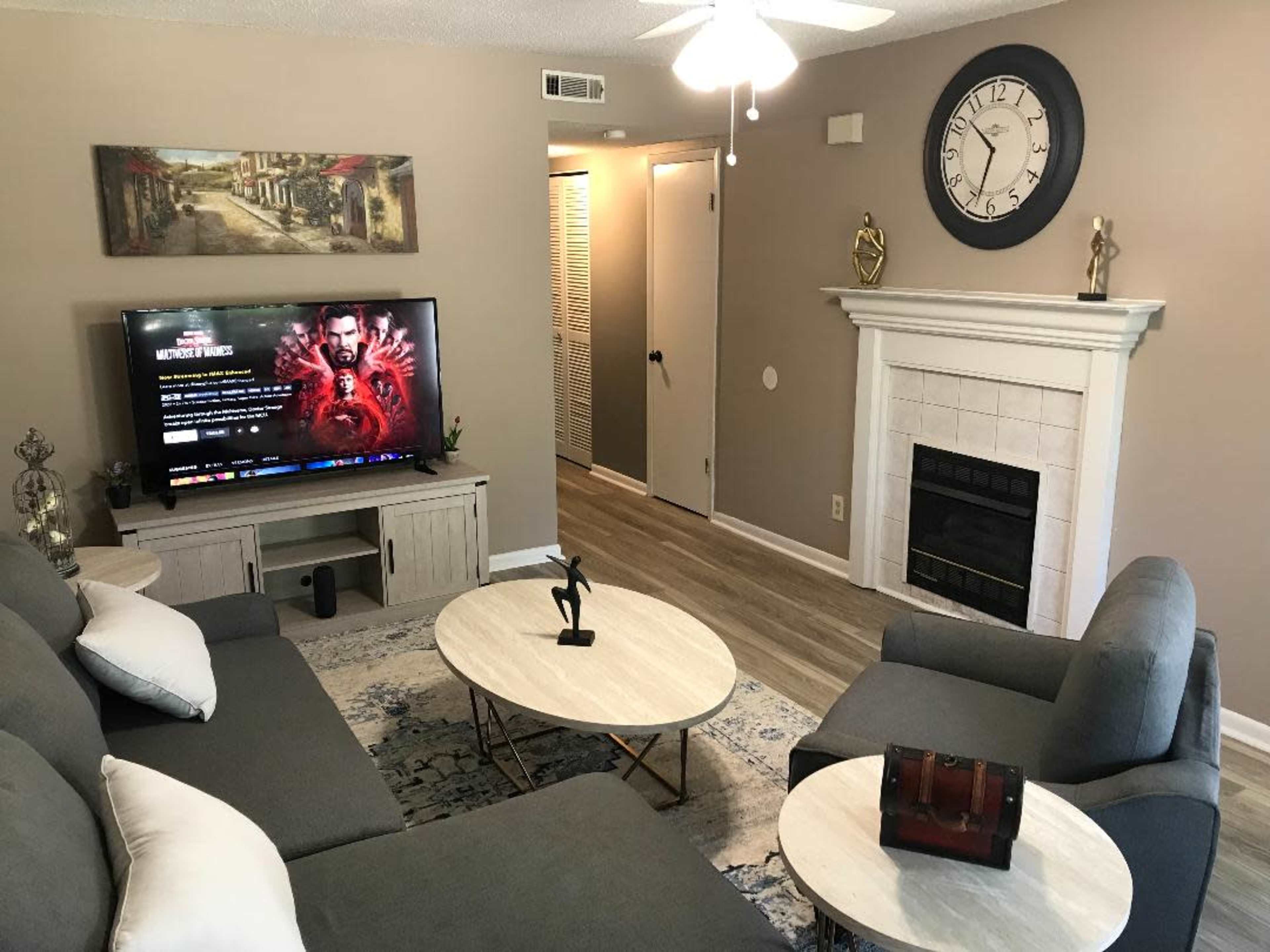 The living room features a television on a stand, a coffee table, two sofas, and a fireplace, with a painting on the wall and a clock above the mantel.