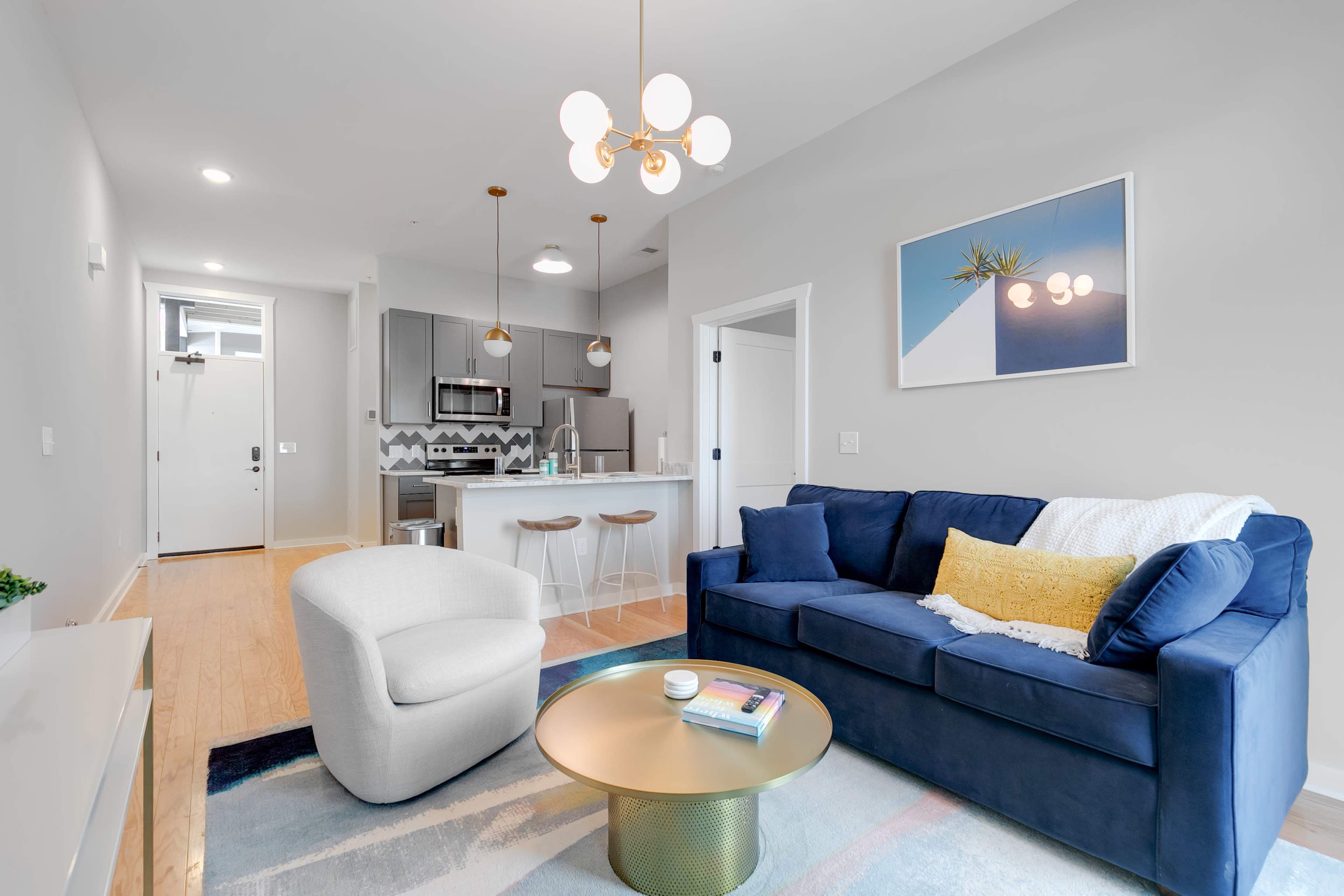 A modern living room with a blue sofa, a white armchair, and a gold coffee table, adjacent to a kitchen area with gray cabinets and stainless steel appliances.