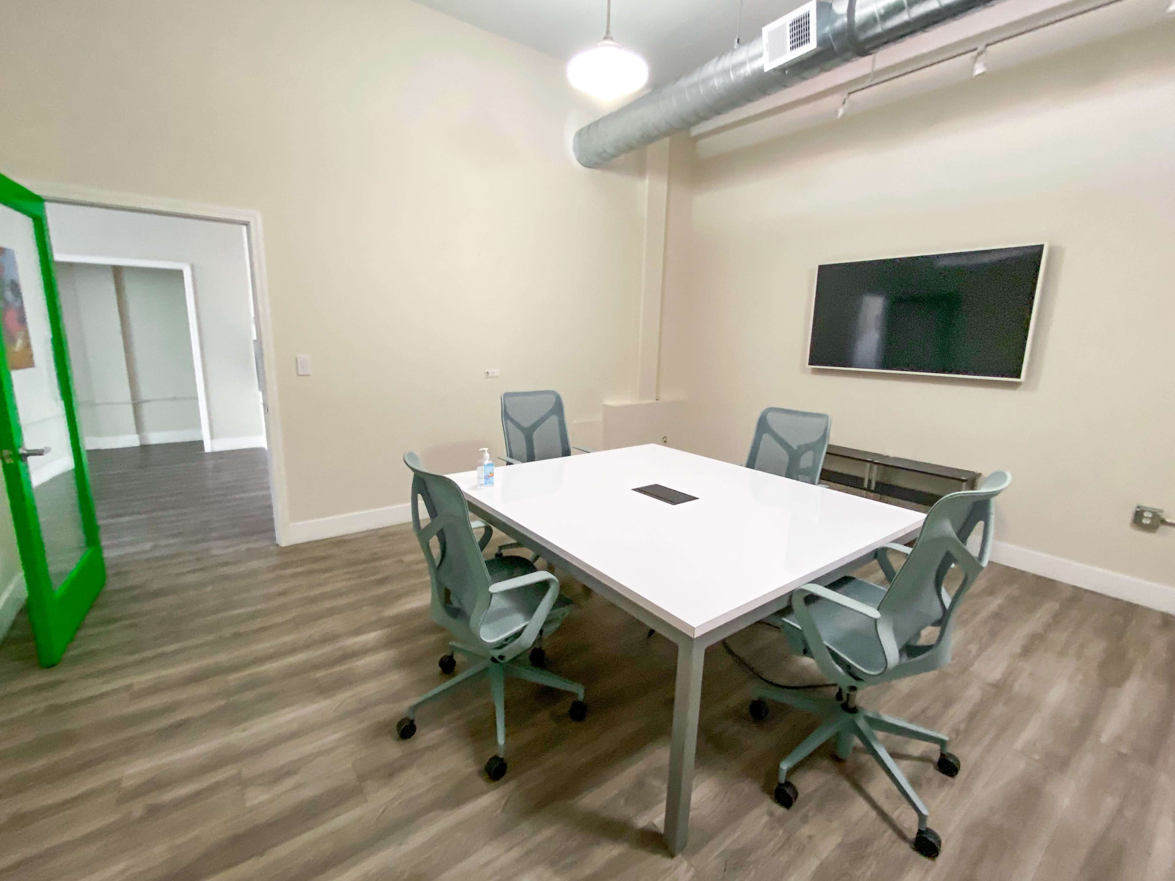 A modern conference room features a white table surrounded by four green chairs, with a television mounted on the wall.