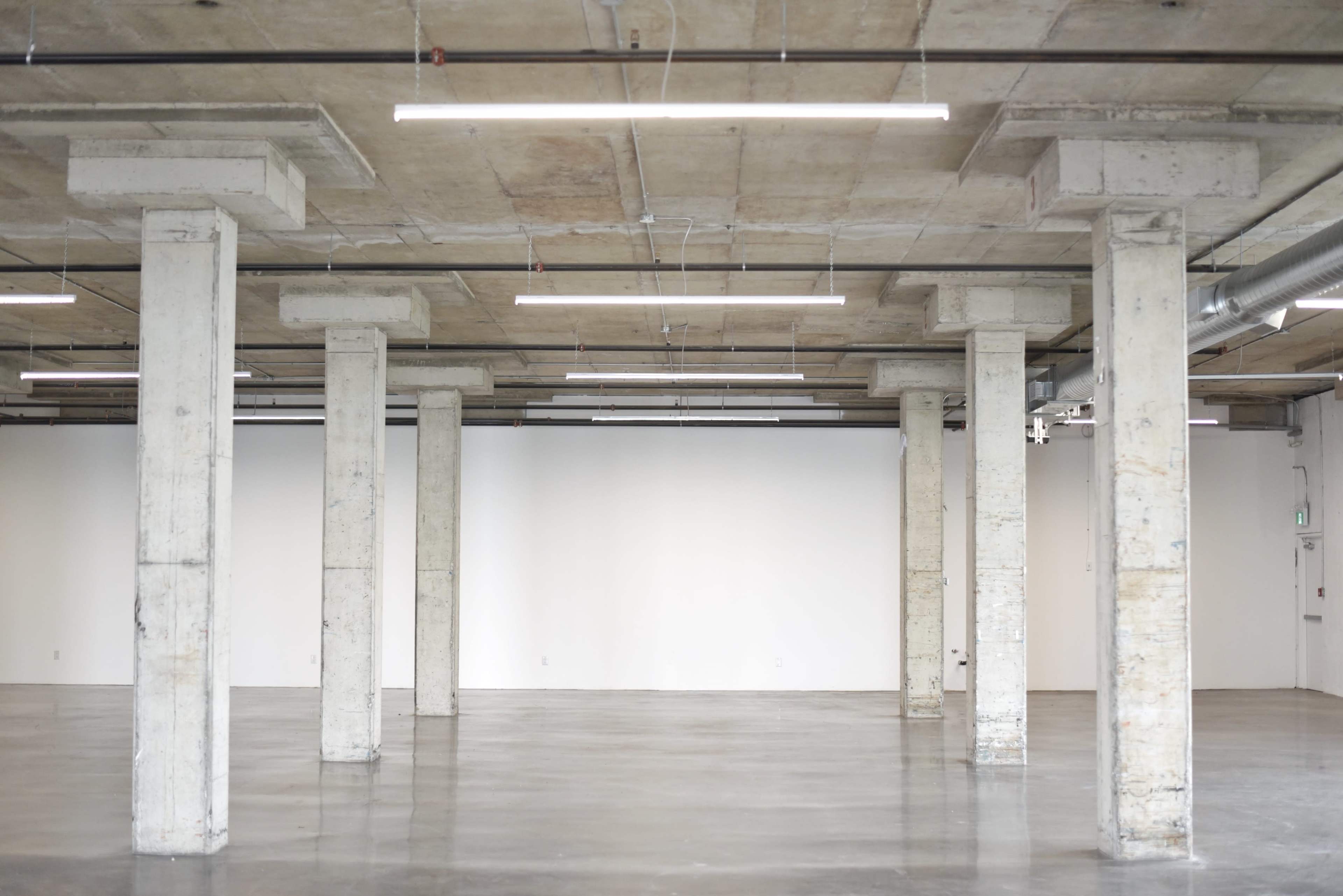 The image shows an empty industrial-style space with concrete pillars and a polished floor, illuminated by overhead lights.
