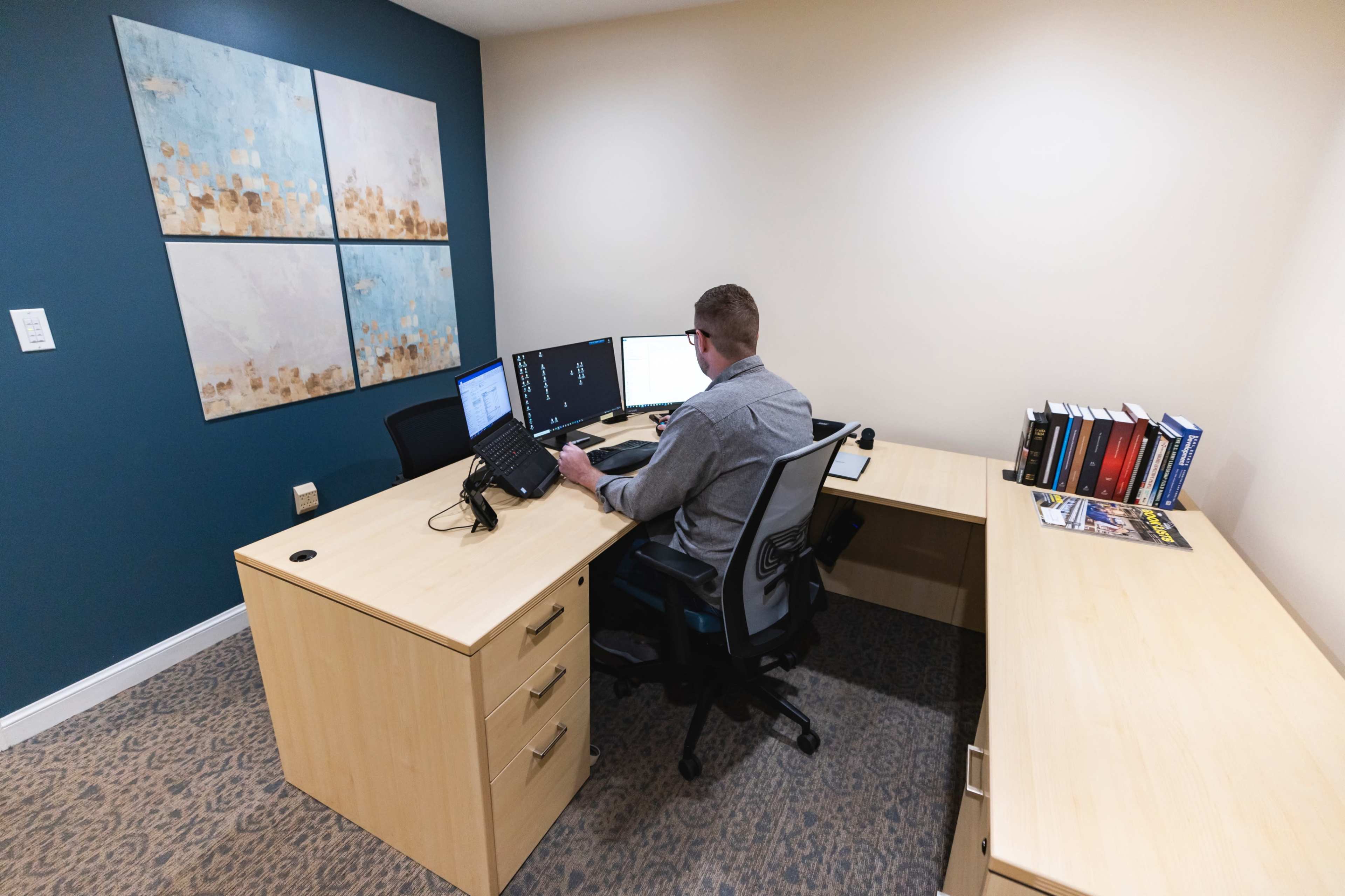 A person sits at a wooden desk in an office, working on dual computer monitors with books lined up on a shelf nearby.