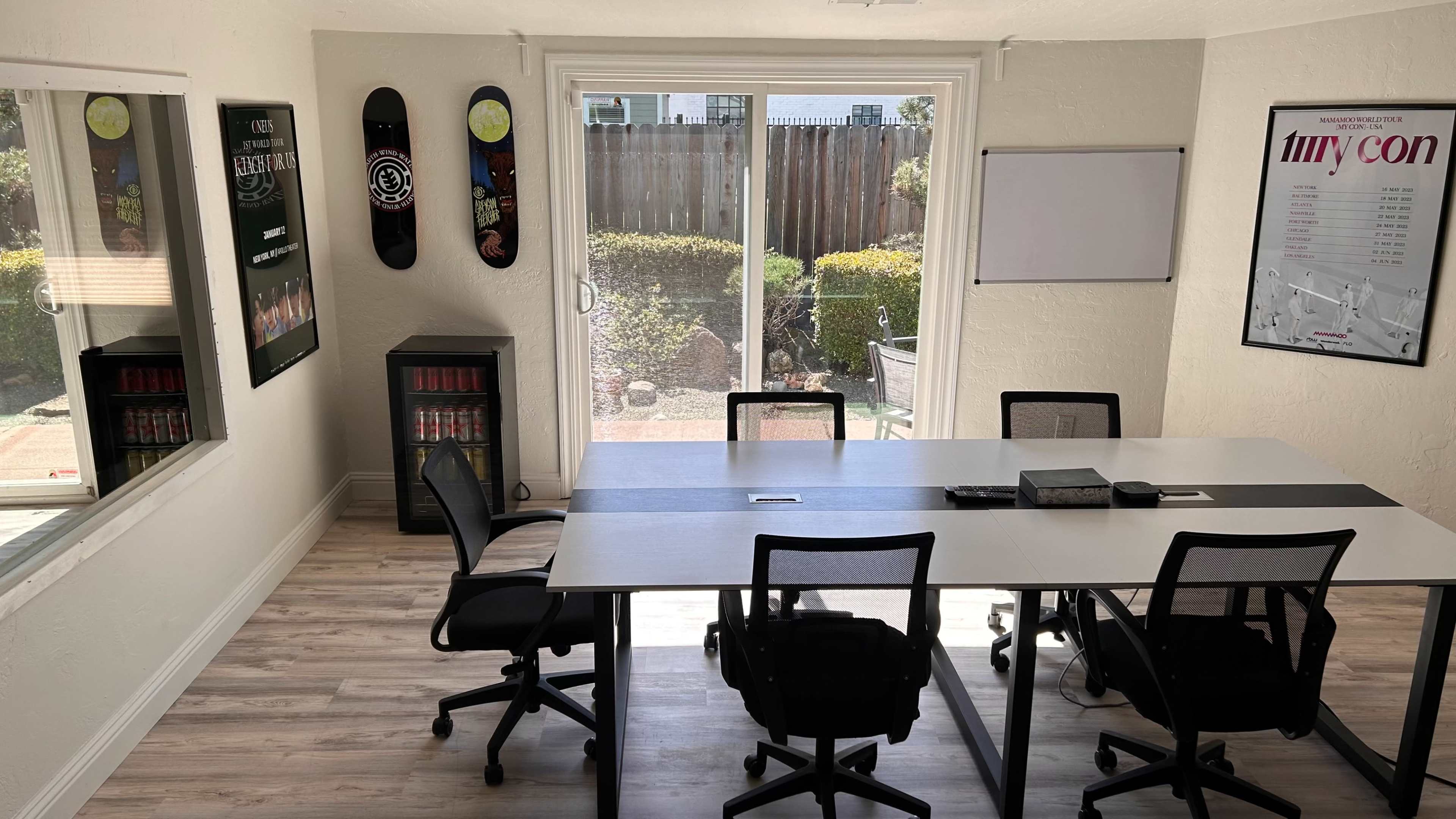 A bright meeting room features a large table surrounded by black chairs, with windows overlooking a garden and posters on the walls.