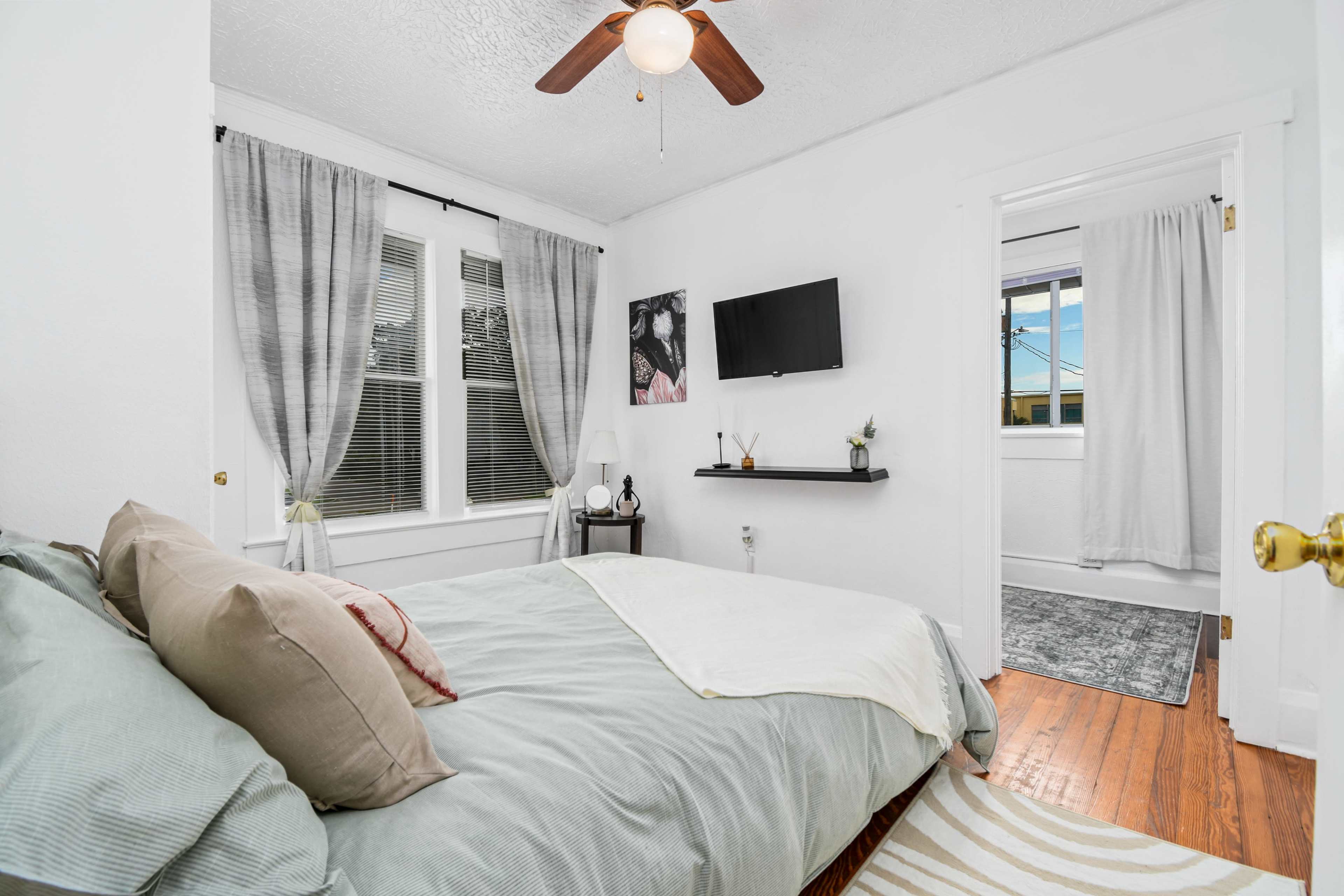 A small, well-lit bedroom featuring a bed with light bedding, a mounted TV on the wall, and a window with sheer curtains.