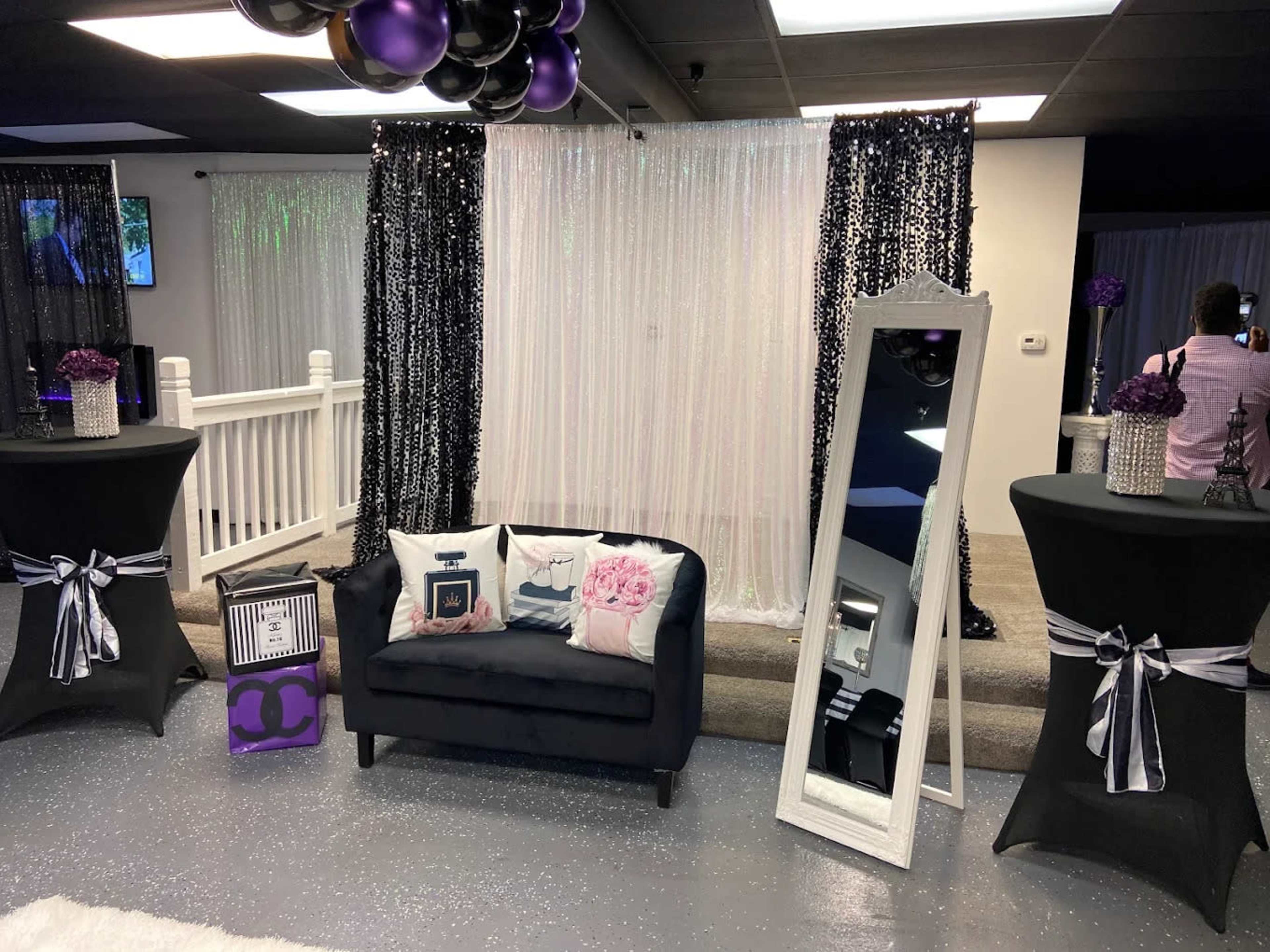 The image shows a decorated event space featuring a black couch, a large mirror, and tables with purple and black accents, surrounded by sequined curtains.