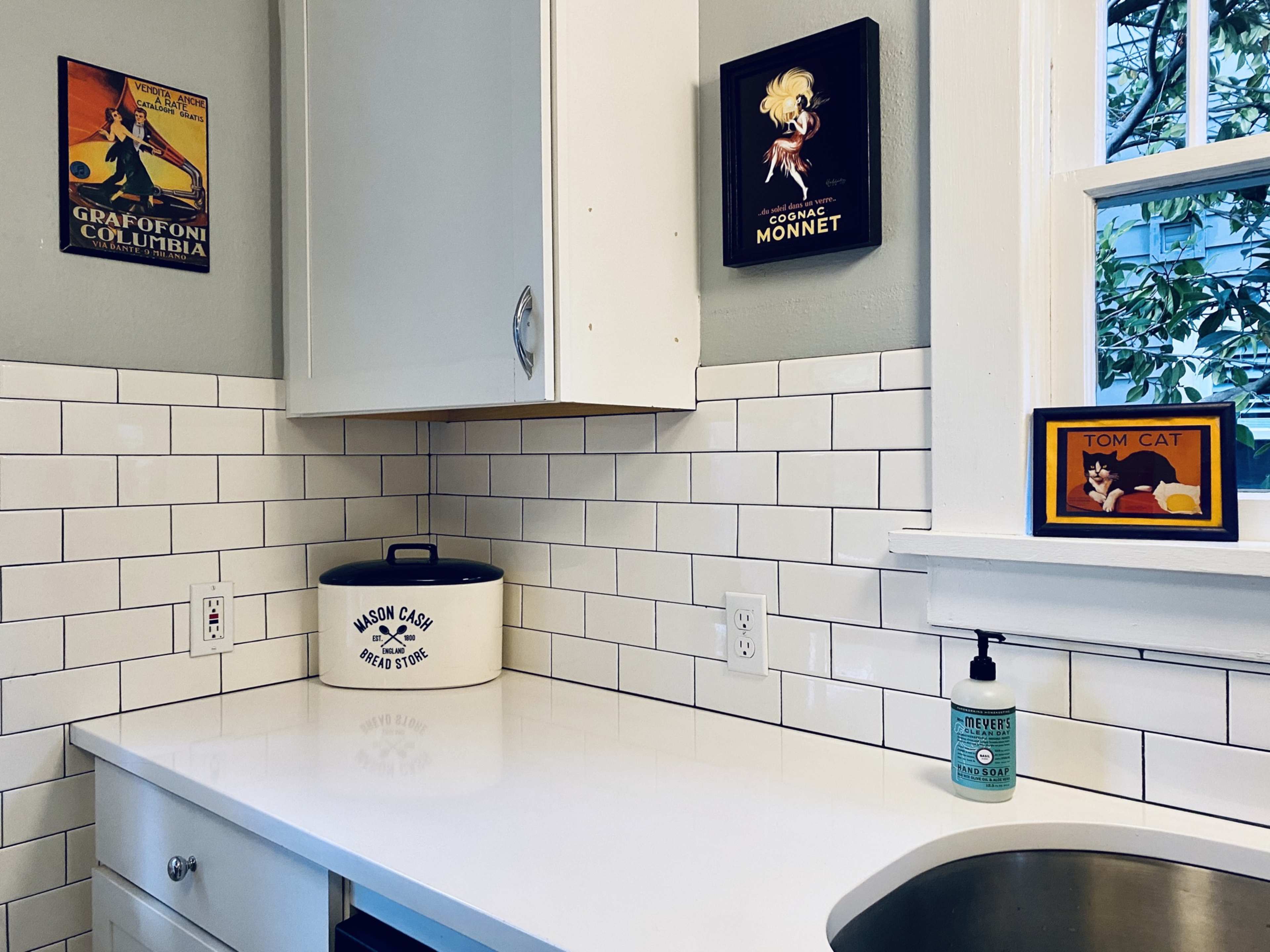 The image shows a kitchen corner with white tiled walls, a countertop, various framed art pieces, a black and white canister, and a hand soap dispenser.