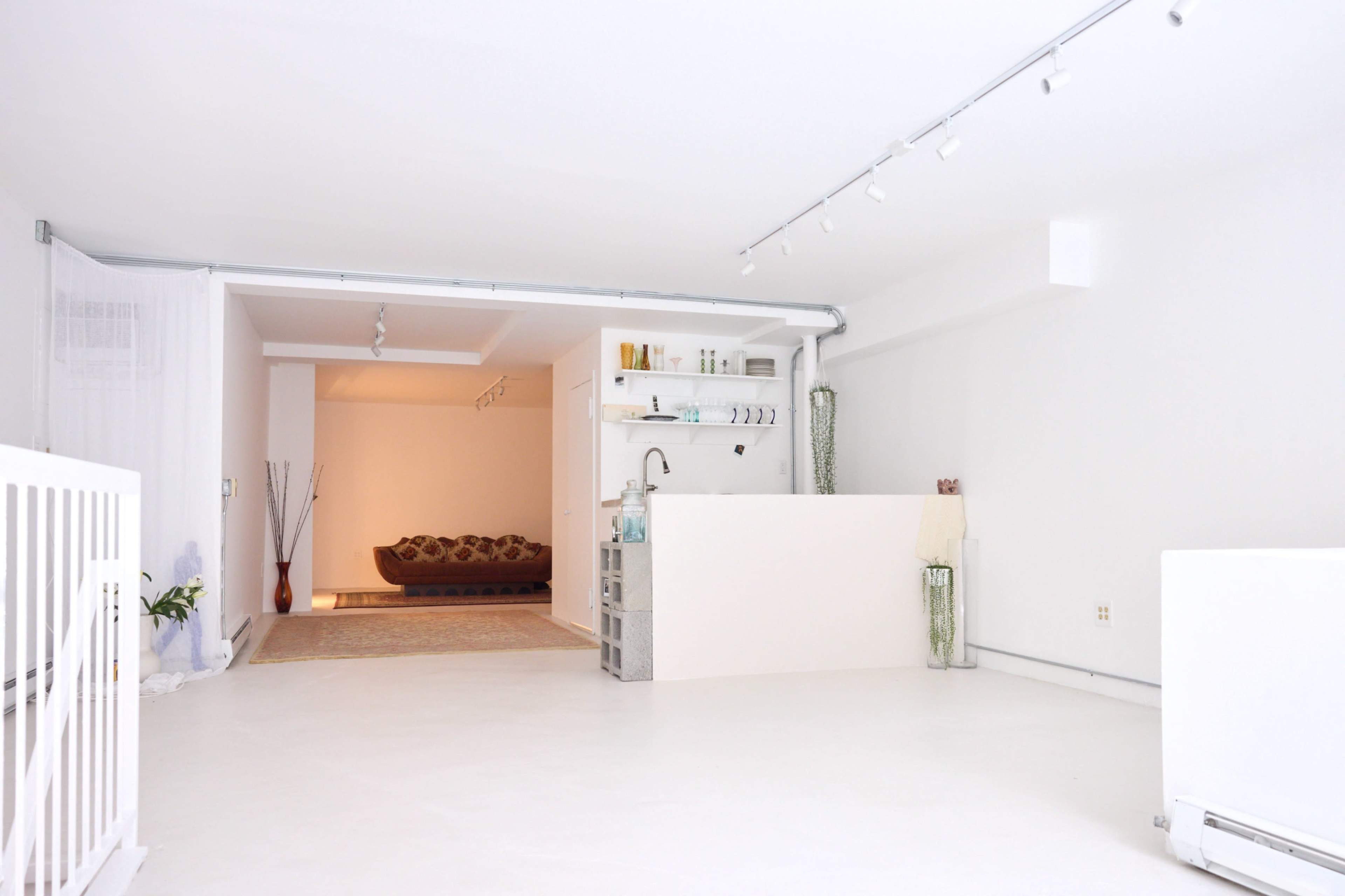 A spacious, minimalist white interior with a small kitchenette and a living area visible in the background.