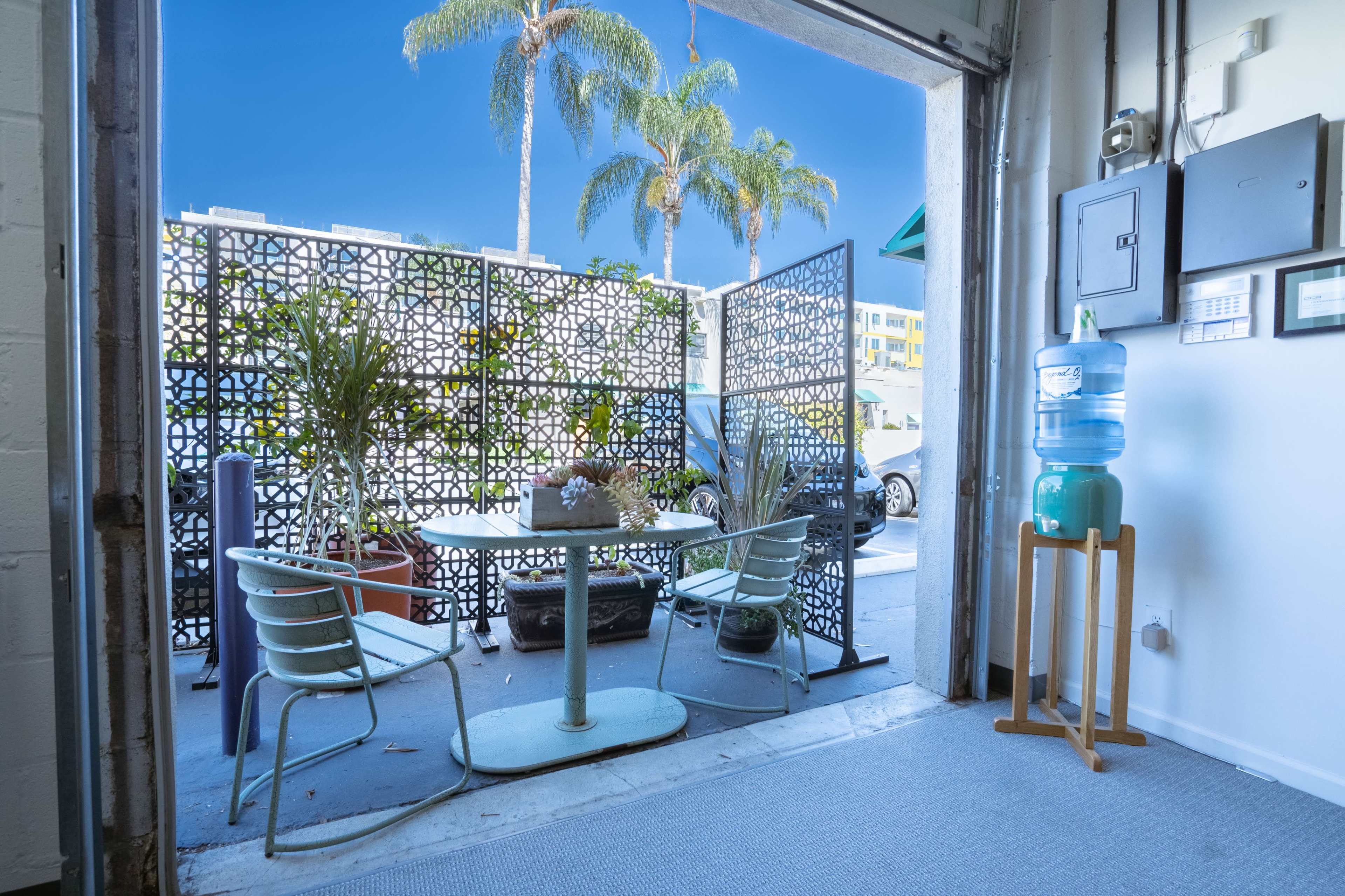 A table and chairs are set up near an open garage door, with a decorative screen and a water cooler nearby, and palm trees visible outside.