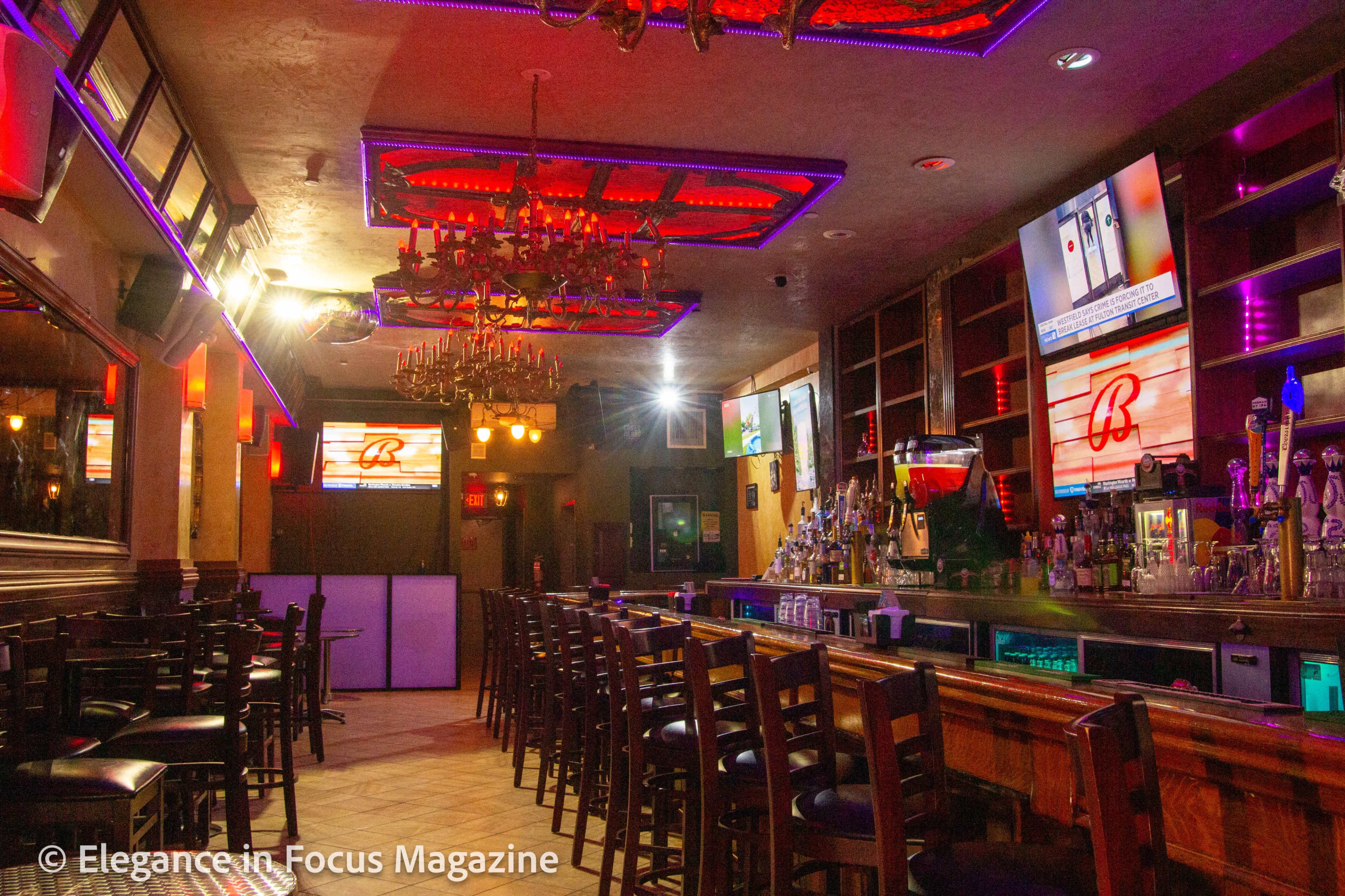 A dimly lit bar with a long counter, multiple screens displaying content, and decorative ceiling lights.