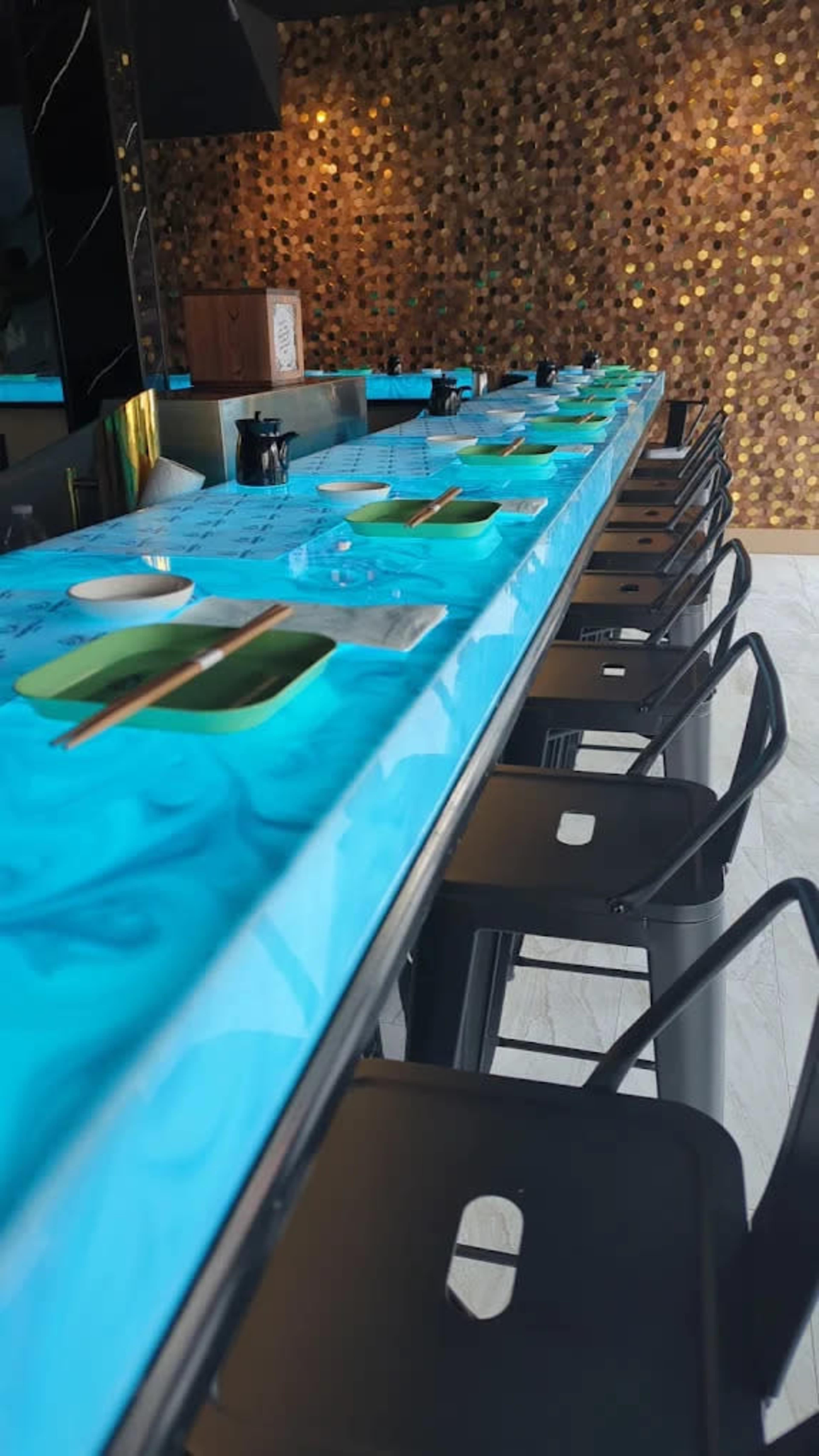 The image shows a dining area with a long, blue marbled table set for a meal, featuring green plates, utensils, and empty chairs lined along one side.