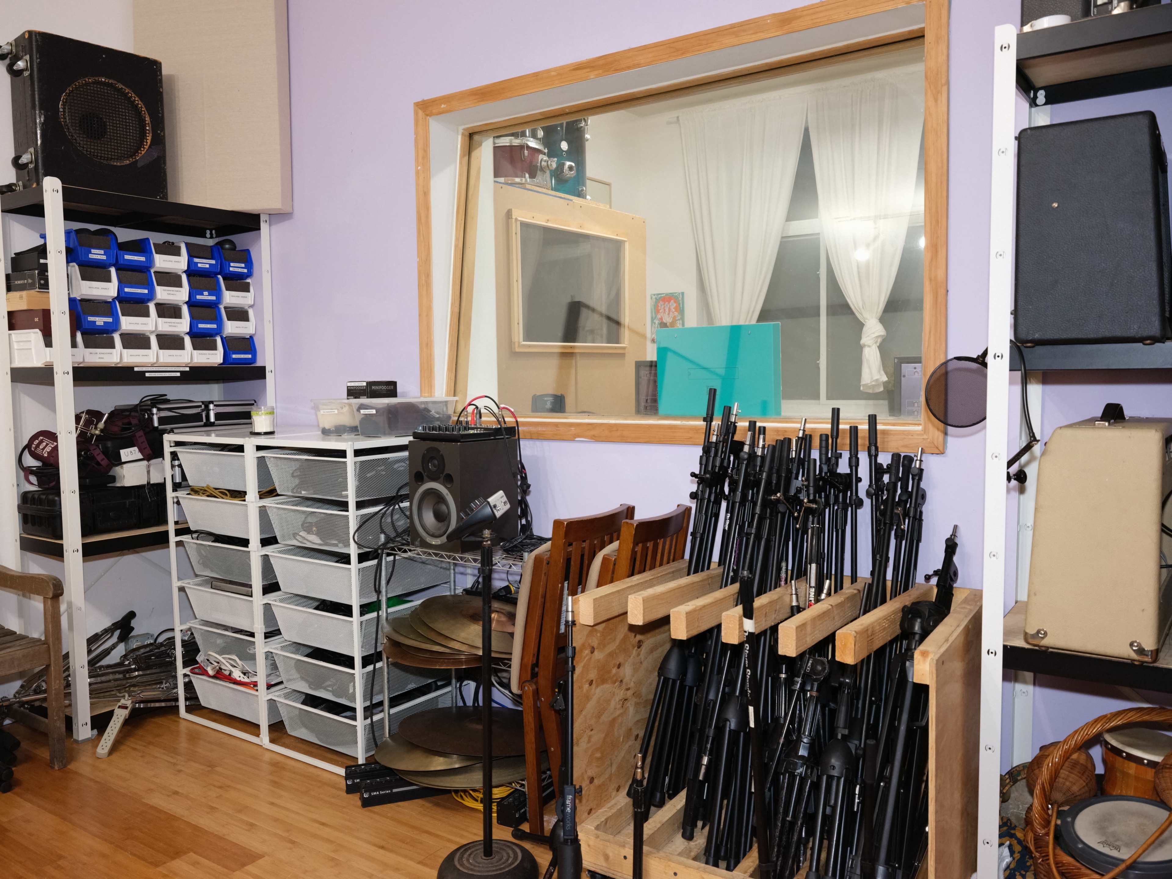 A recording studio with a glass window, shelves filled with equipment, and a collection of microphone stands stacked in a corner.