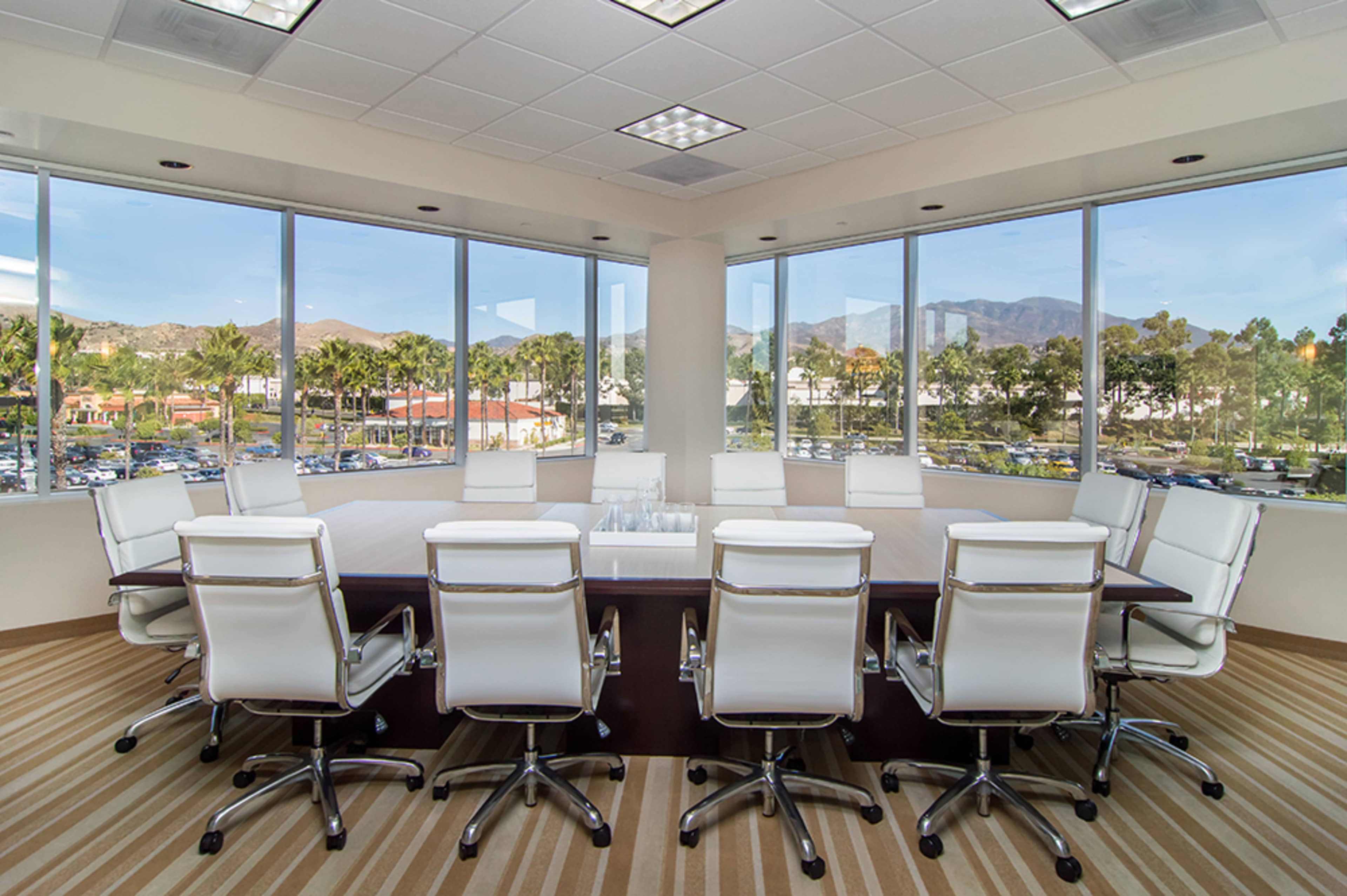 A conference room with a large table surrounded by white chairs and windows offering a view of mountains and palm trees outside.