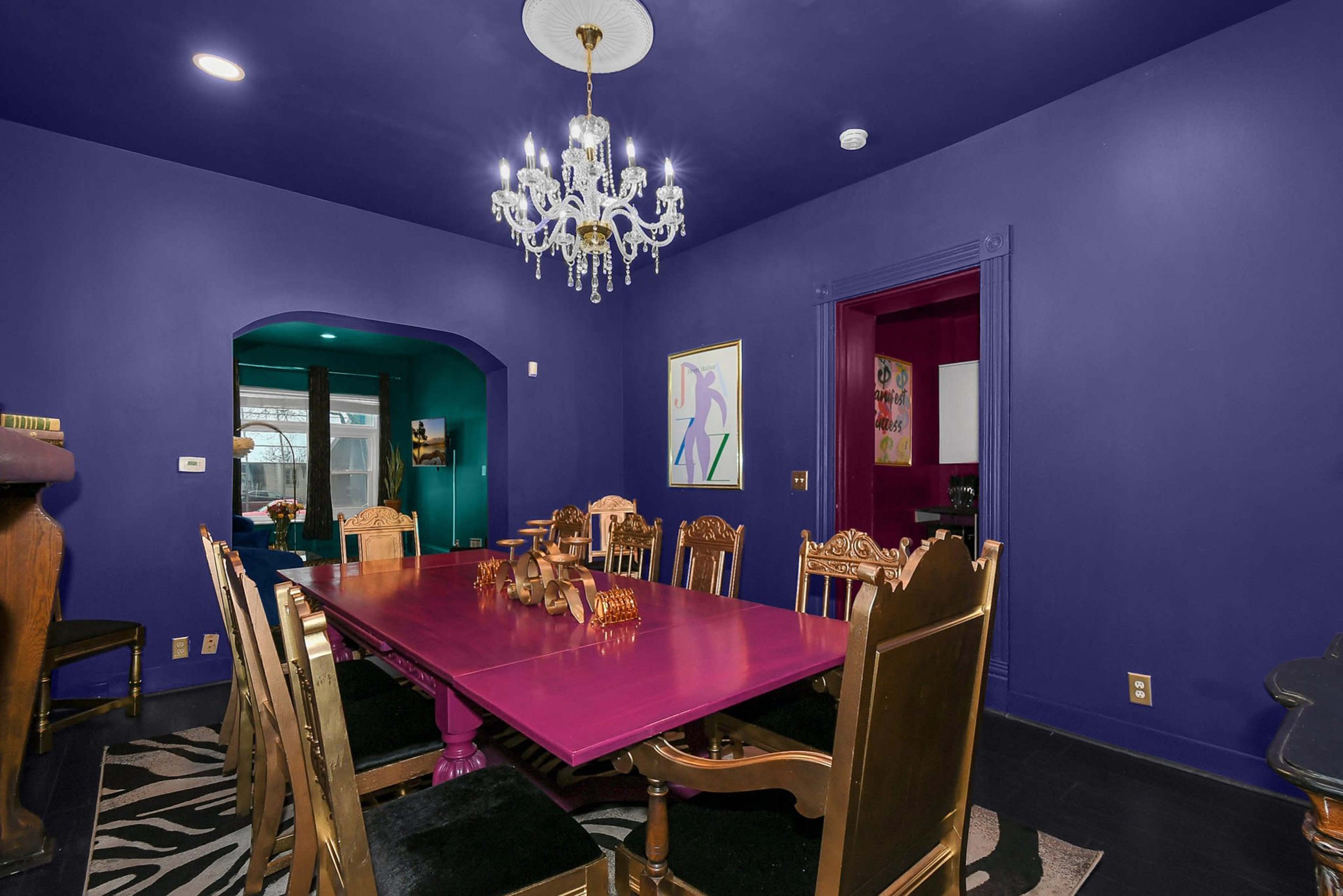 The dining room features a large purple table surrounded by ornate wooden chairs, with a chandelier hanging above and dark purple walls.