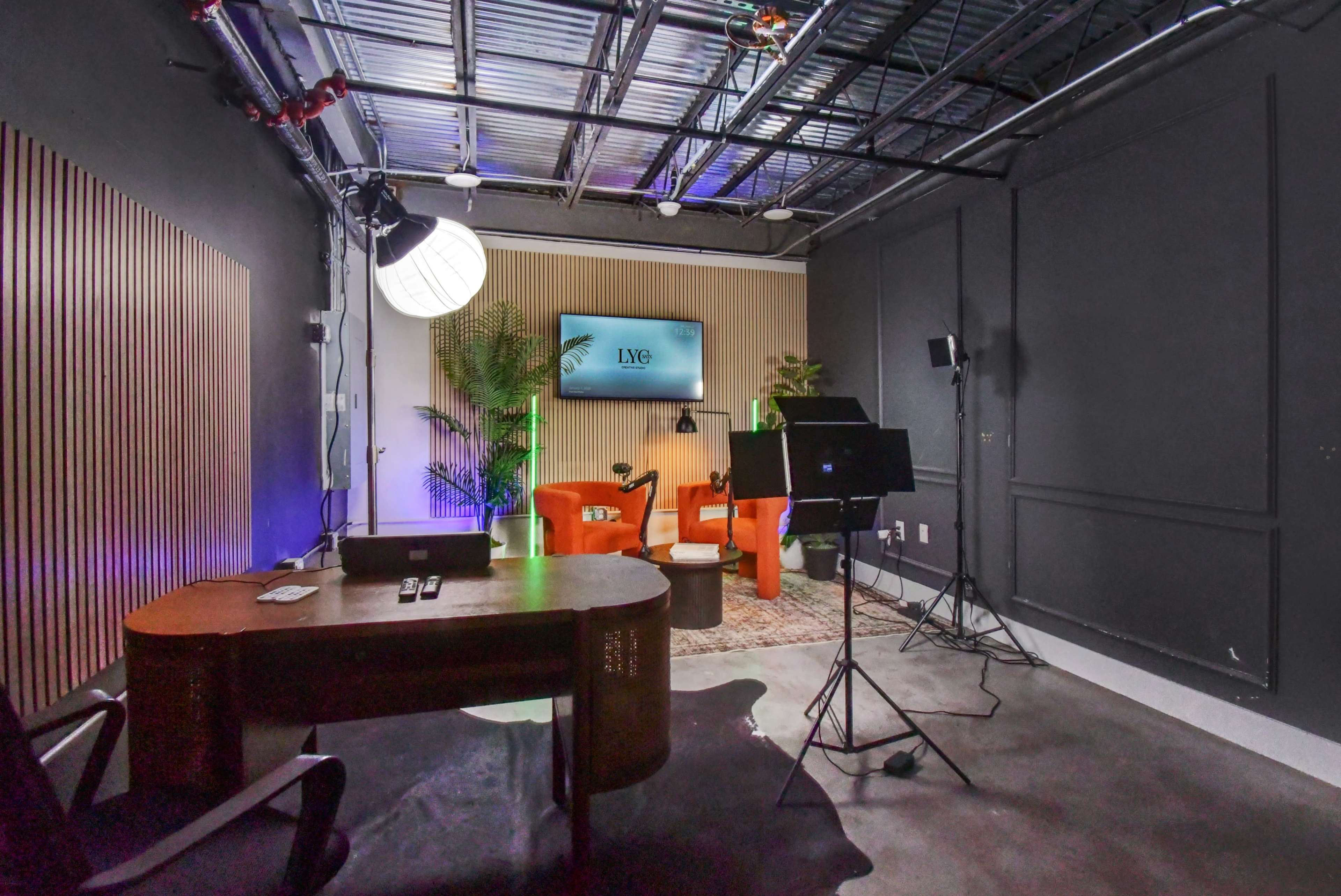 The image shows a modern studio space featuring a desk, two orange chairs, a television on the wall, and various lighting and filming equipment.