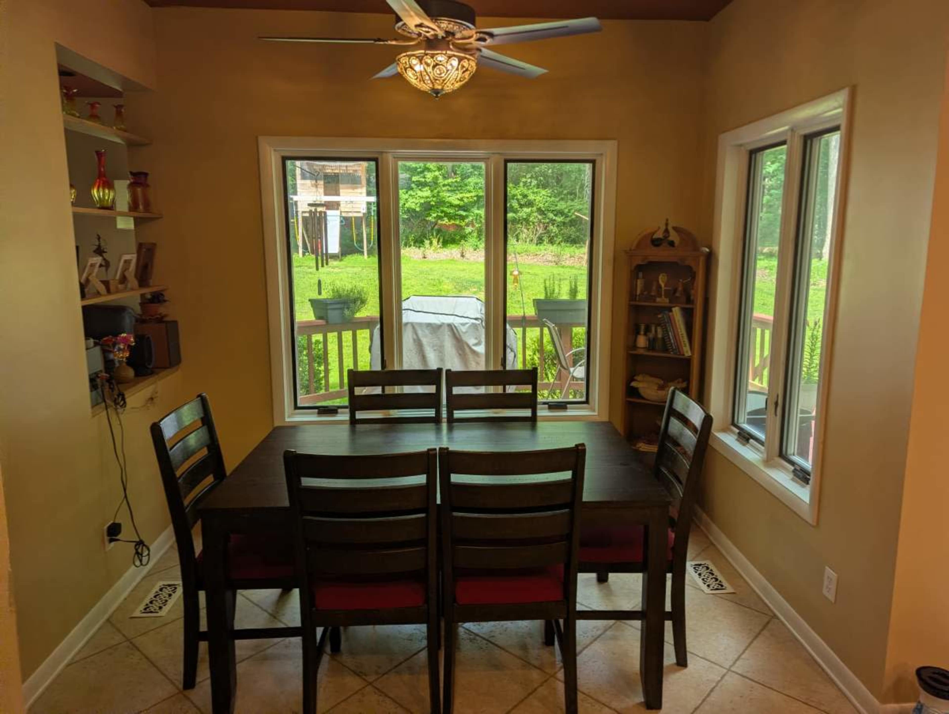 A wooden dining table with six chairs is set in a well-lit room with large windows overlooking a backyard.