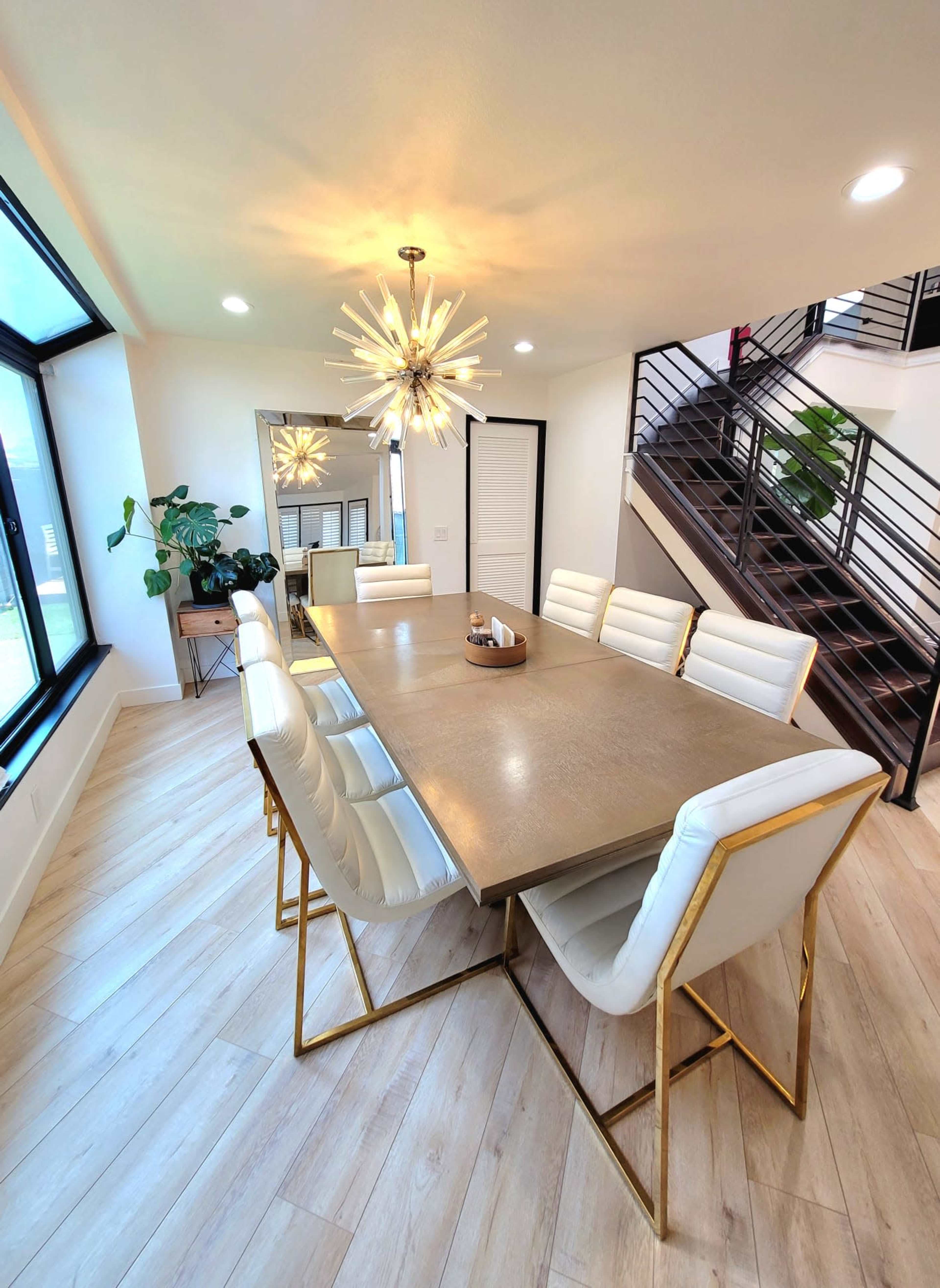 A modern dining area features a large rectangular table surrounded by white upholstered chairs, with a distinctive chandelier overhead and a staircase in the background.
