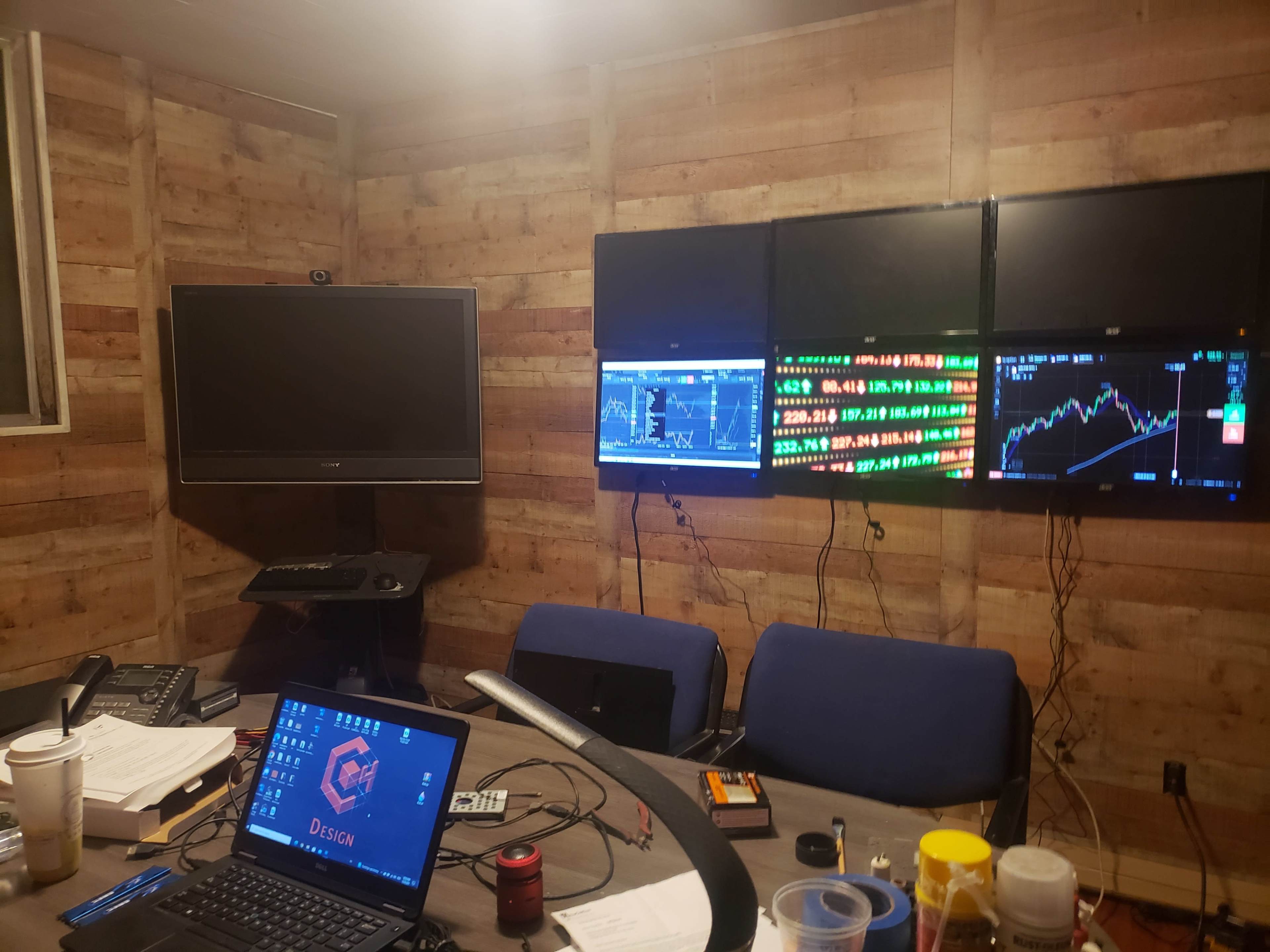 The image shows a dimly lit room with a wooden wall, featuring multiple computer monitors displaying financial charts and data, alongside a desk with a laptop and various office supplies.