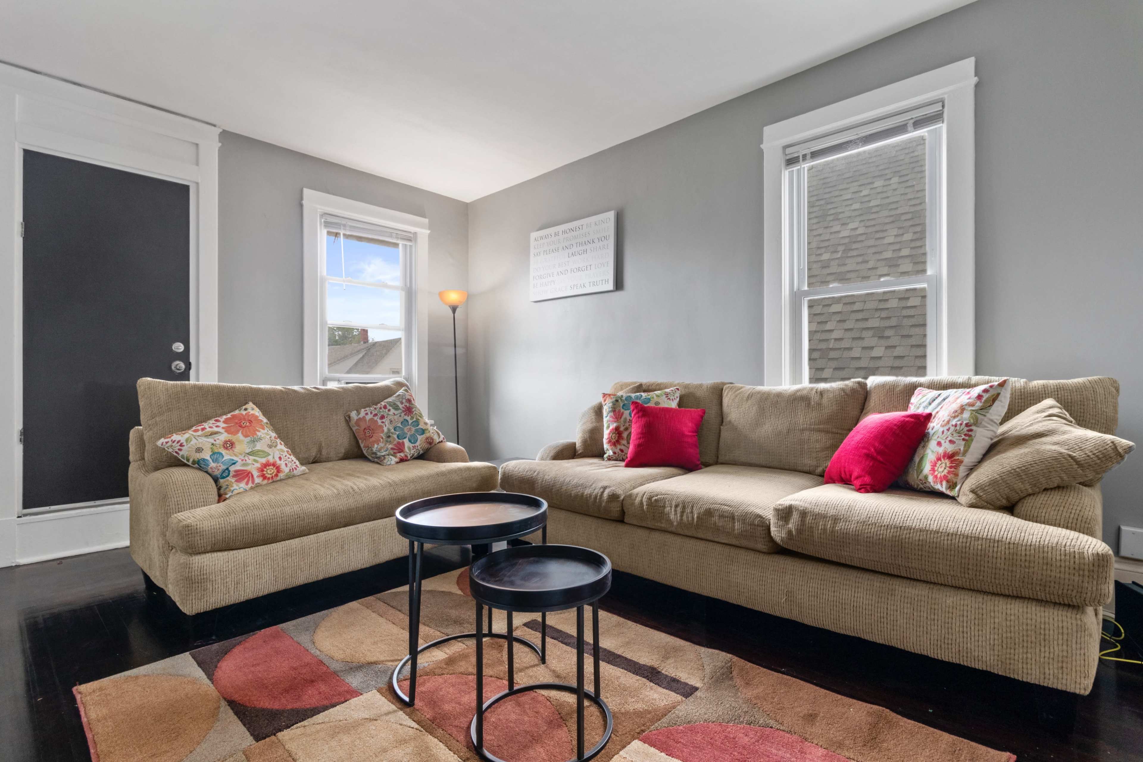 A cozy living room featuring a beige sofa with colorful cushions, two small black tables, and large windows allowing natural light.