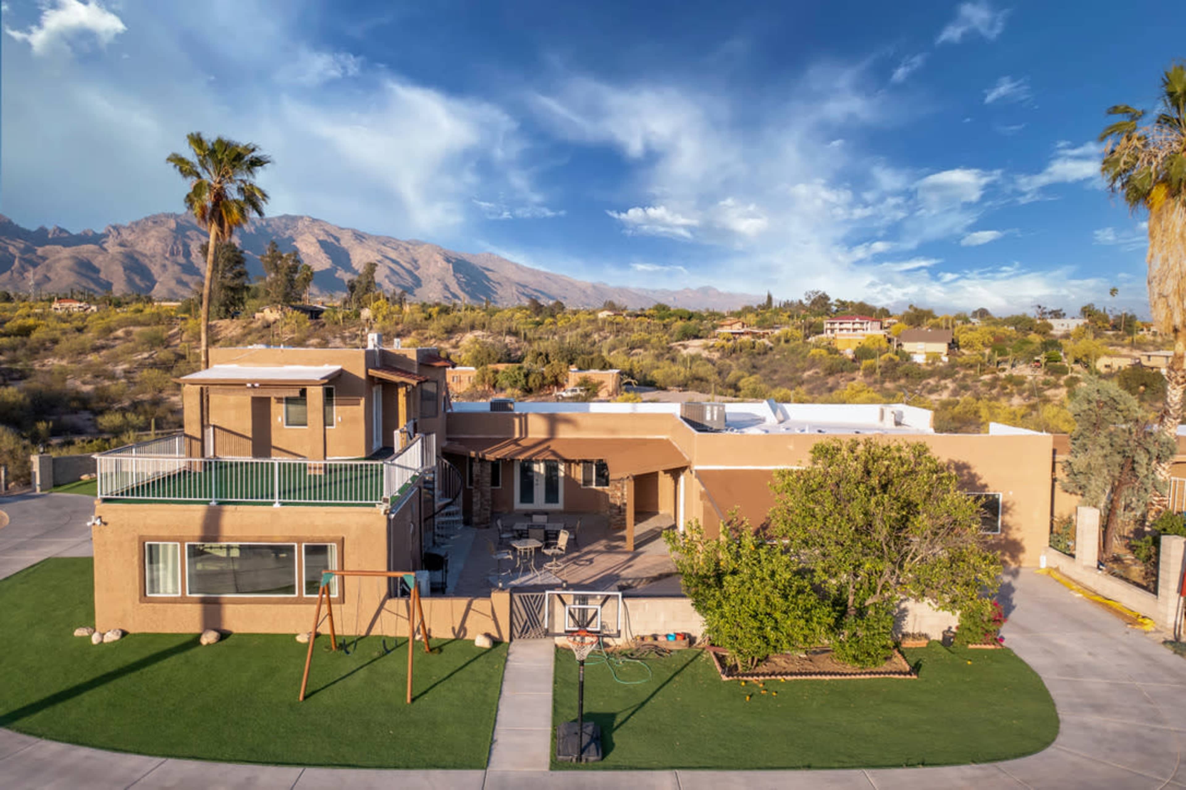 A large tan house with a patio and a swing set in the front yard, surrounded by mountains and palm trees.