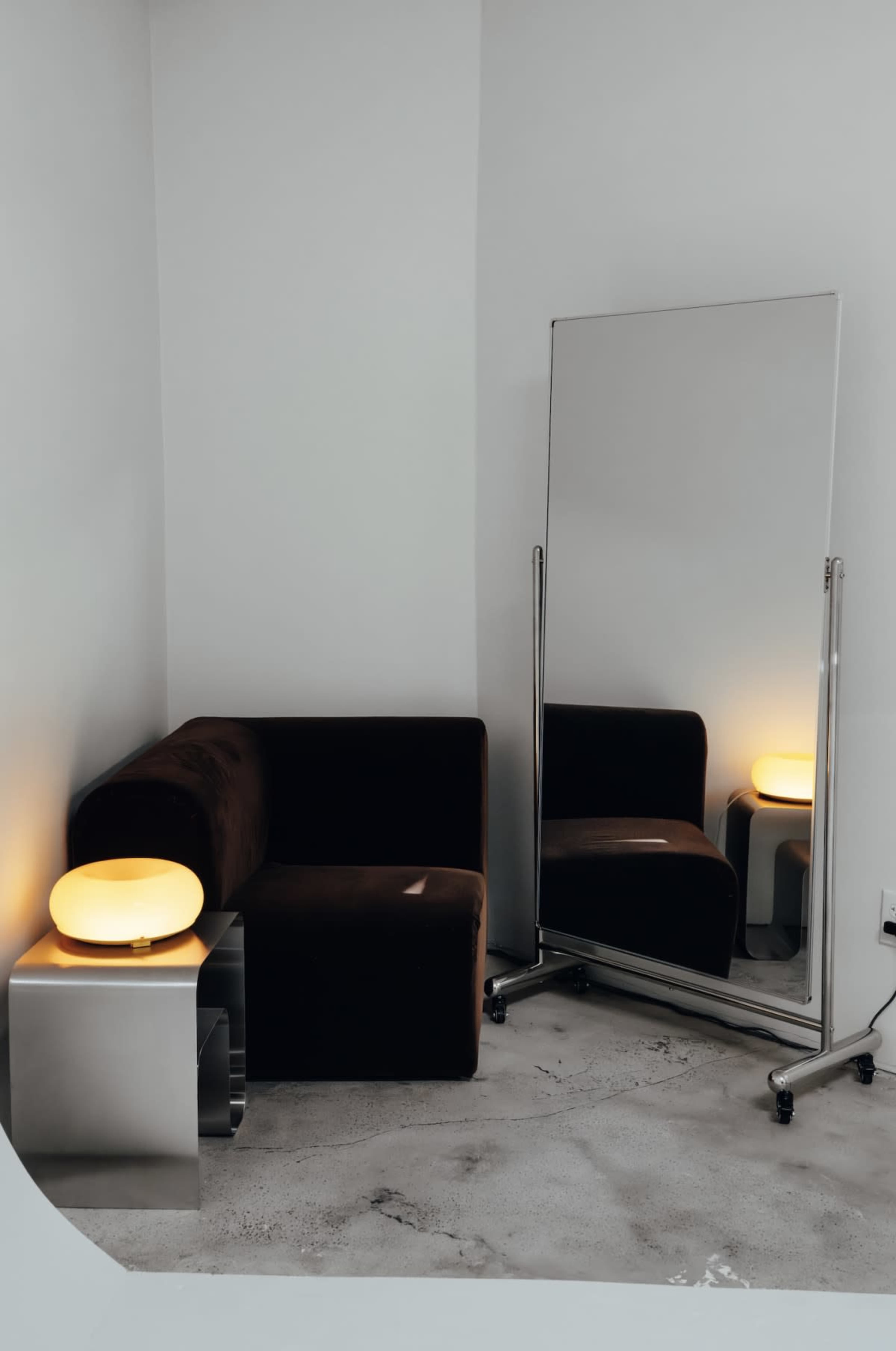 The image shows a minimalist room featuring a dark couch, a tall mirror on wheels, and a modern lamp on a metal side table.