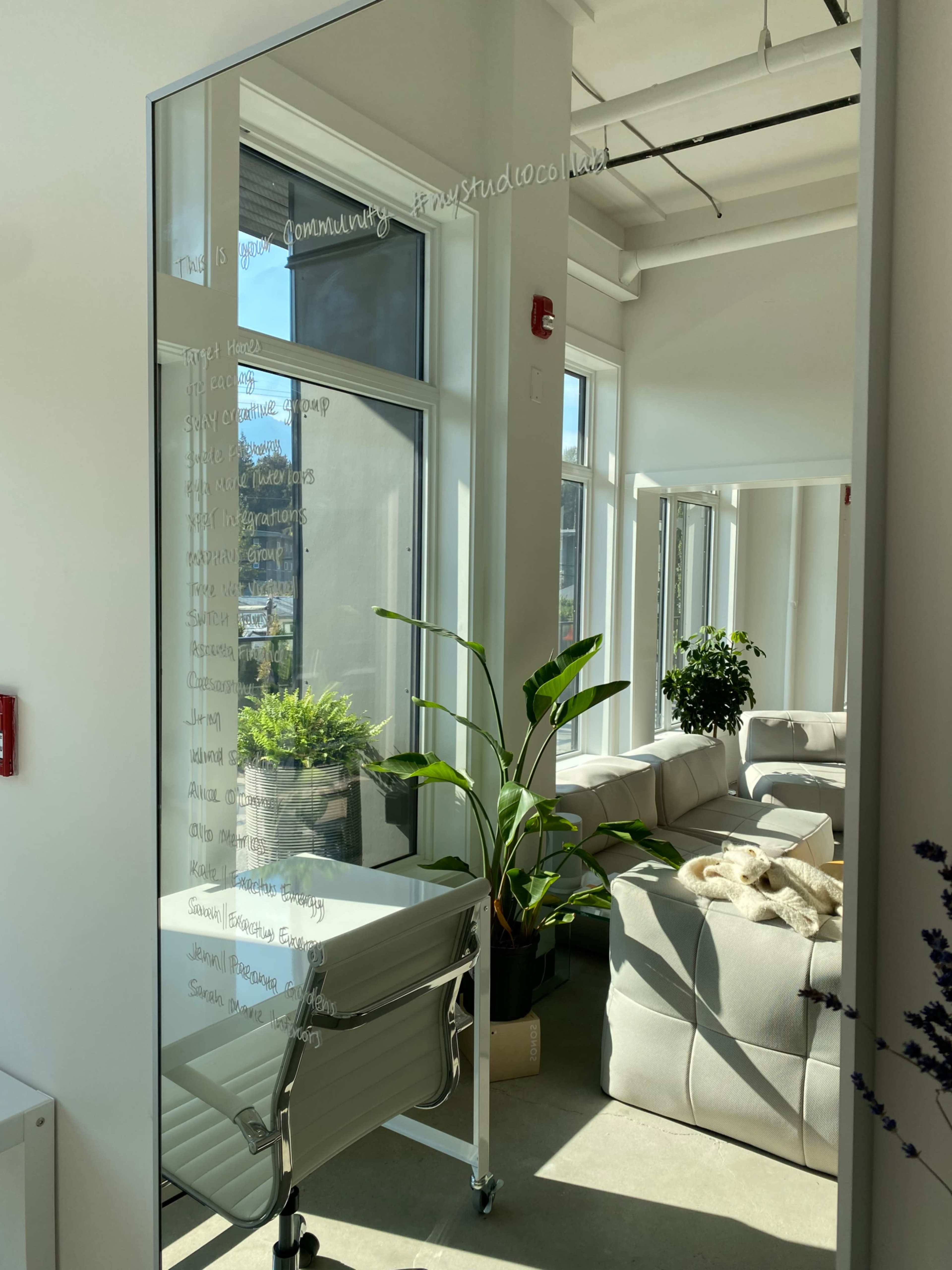 A large mirror reflects a bright office space with a chair and a decorative plant, adjacent to a white sofa and large windows.