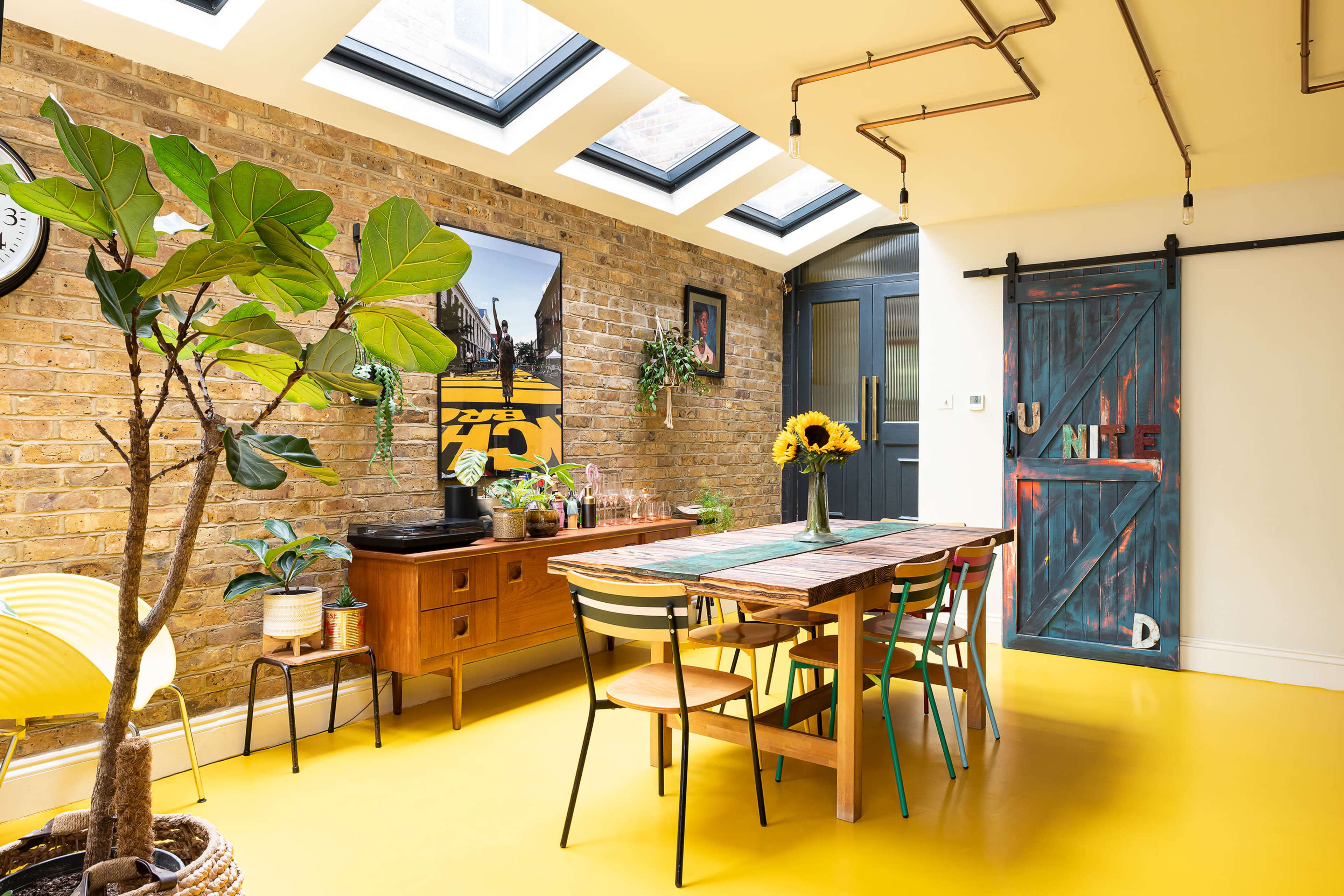 The bright dining area features a long wooden table surrounded by colorful chairs, against a backdrop of exposed brick walls and large skylights.