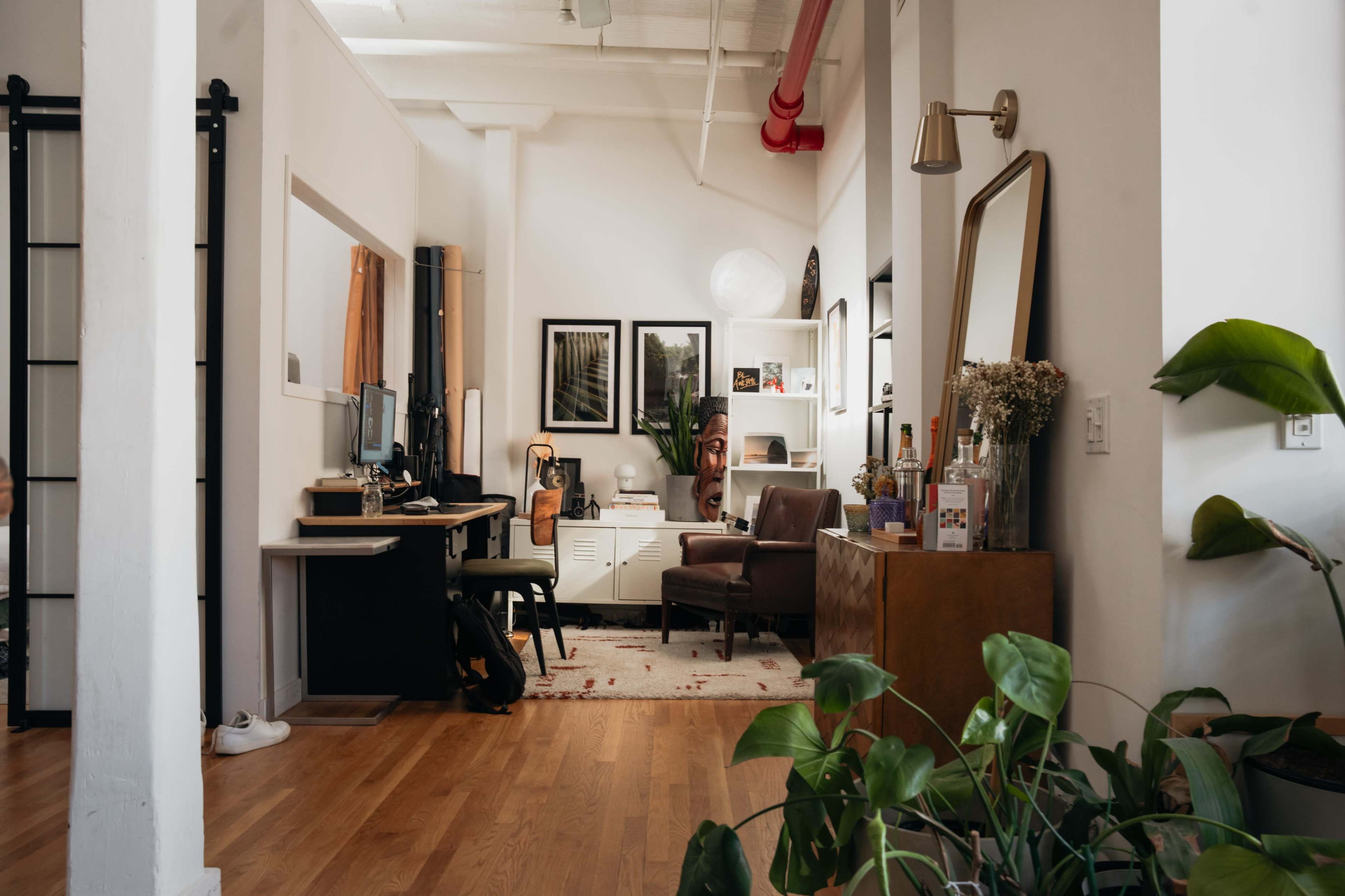 The image shows a modern office space with a wooden floor, desk with a computer, a chair, and decorative shelves featuring artwork and plants.