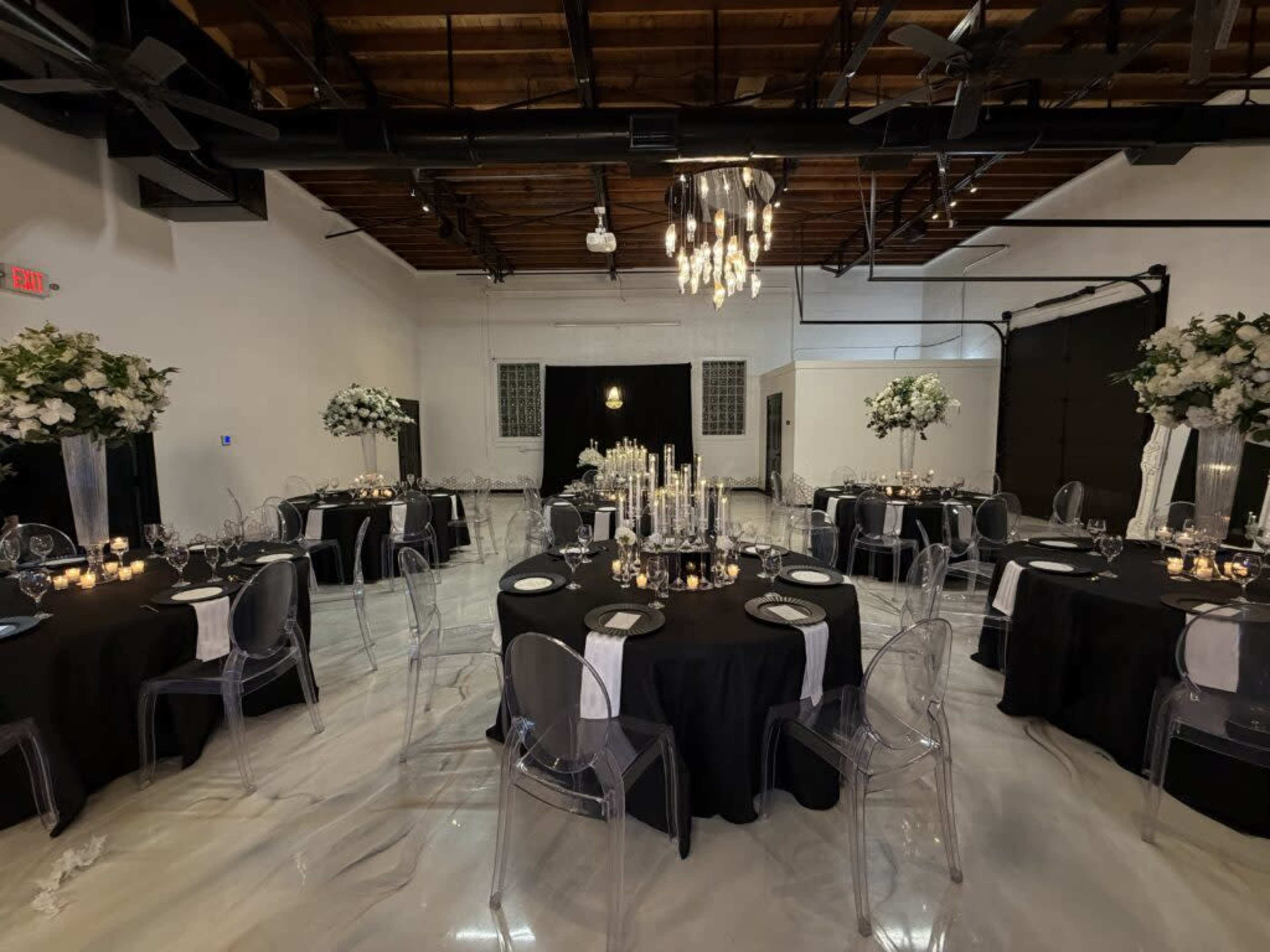 The scene shows a well-decorated event space featuring round tables with black tablecloths, clear ghost chairs, and elegant centerpieces of flowers and candles.