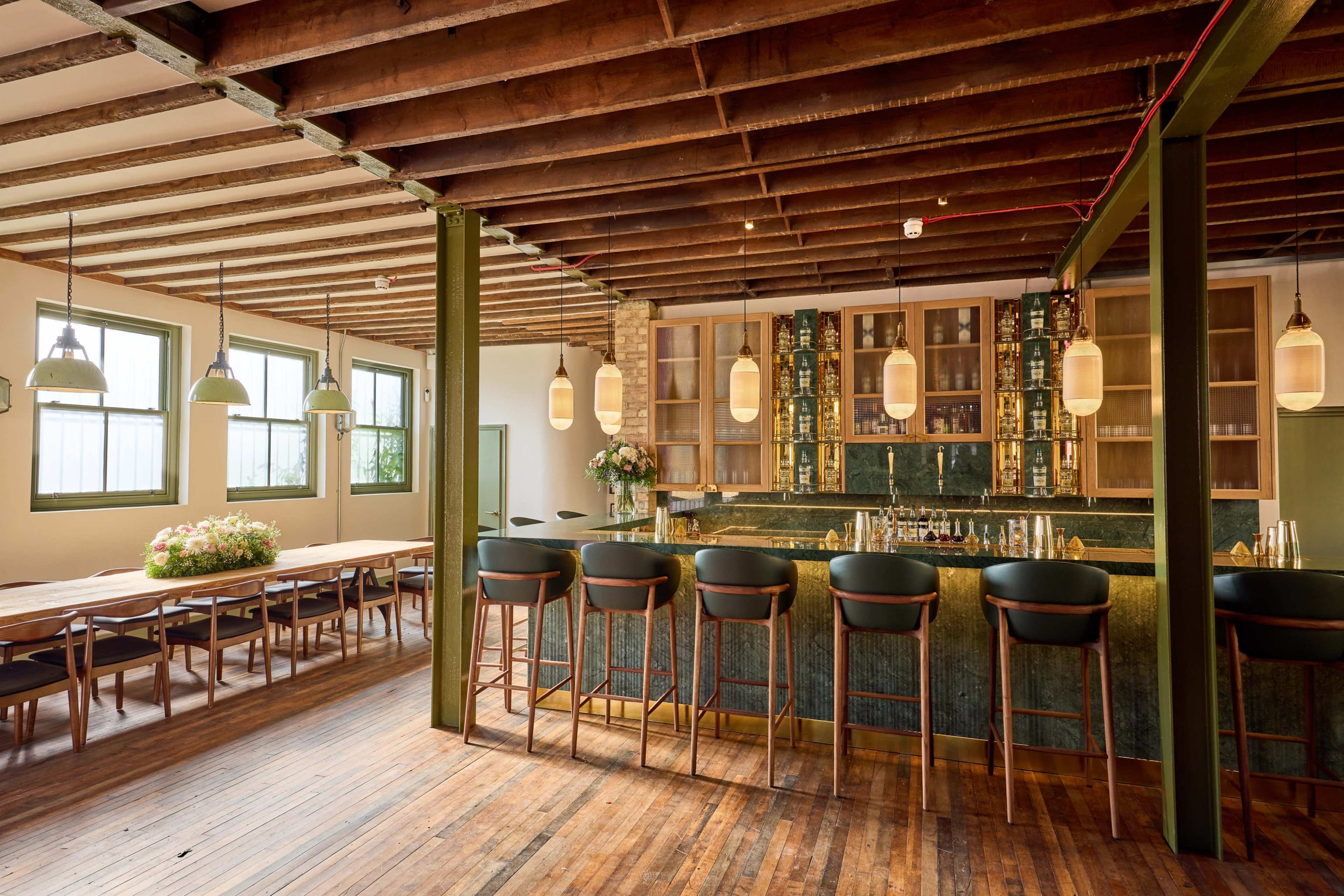 The image shows a modern bar area with a green marble counter, wooden beams on the ceiling, and seating along the bar, adjacent to a dining space with a long wooden table.