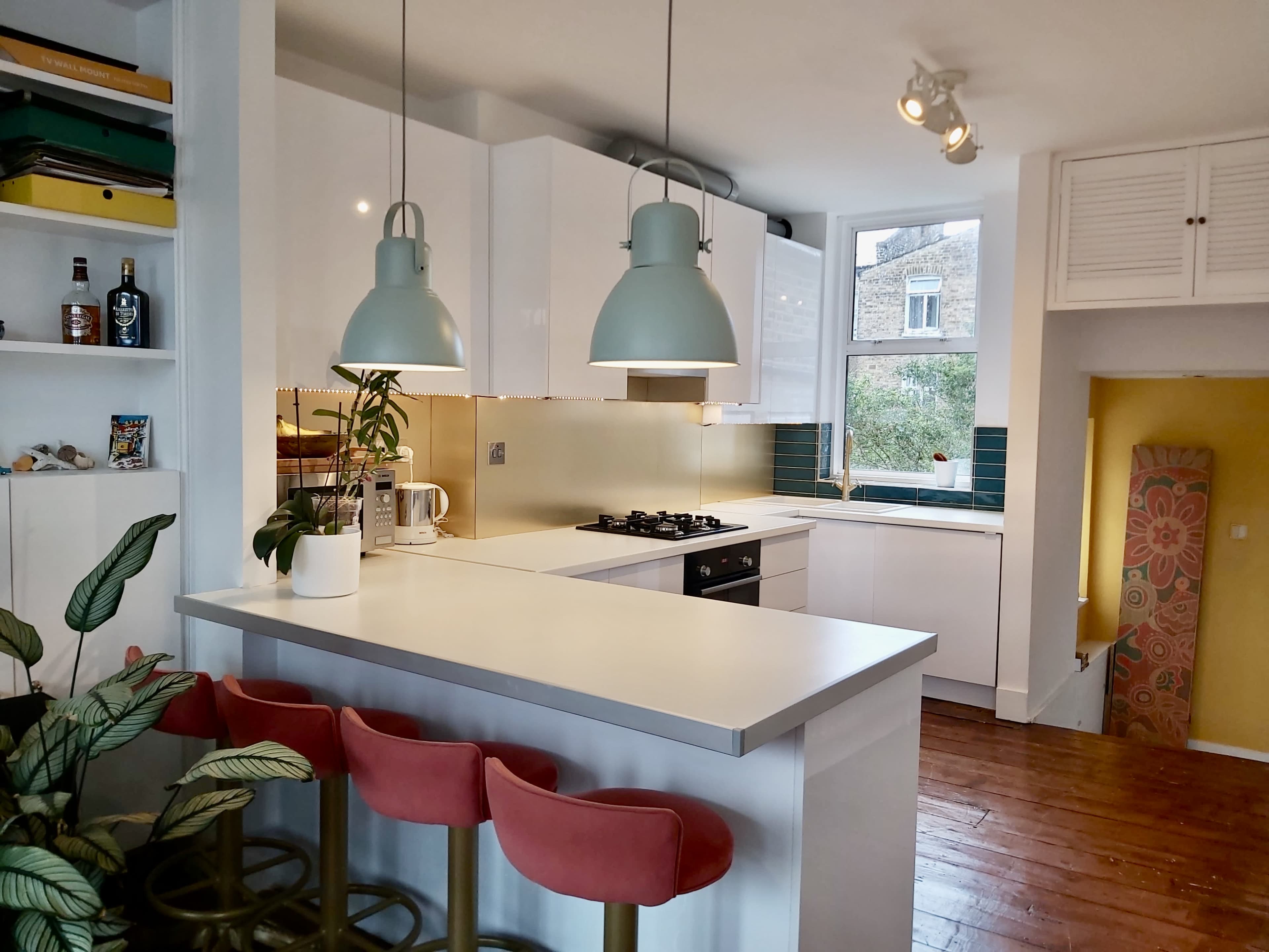 A modern kitchen with white cabinets, a gas stove, and a breakfast bar featuring three pink stools, illuminated by two green pendant lights.