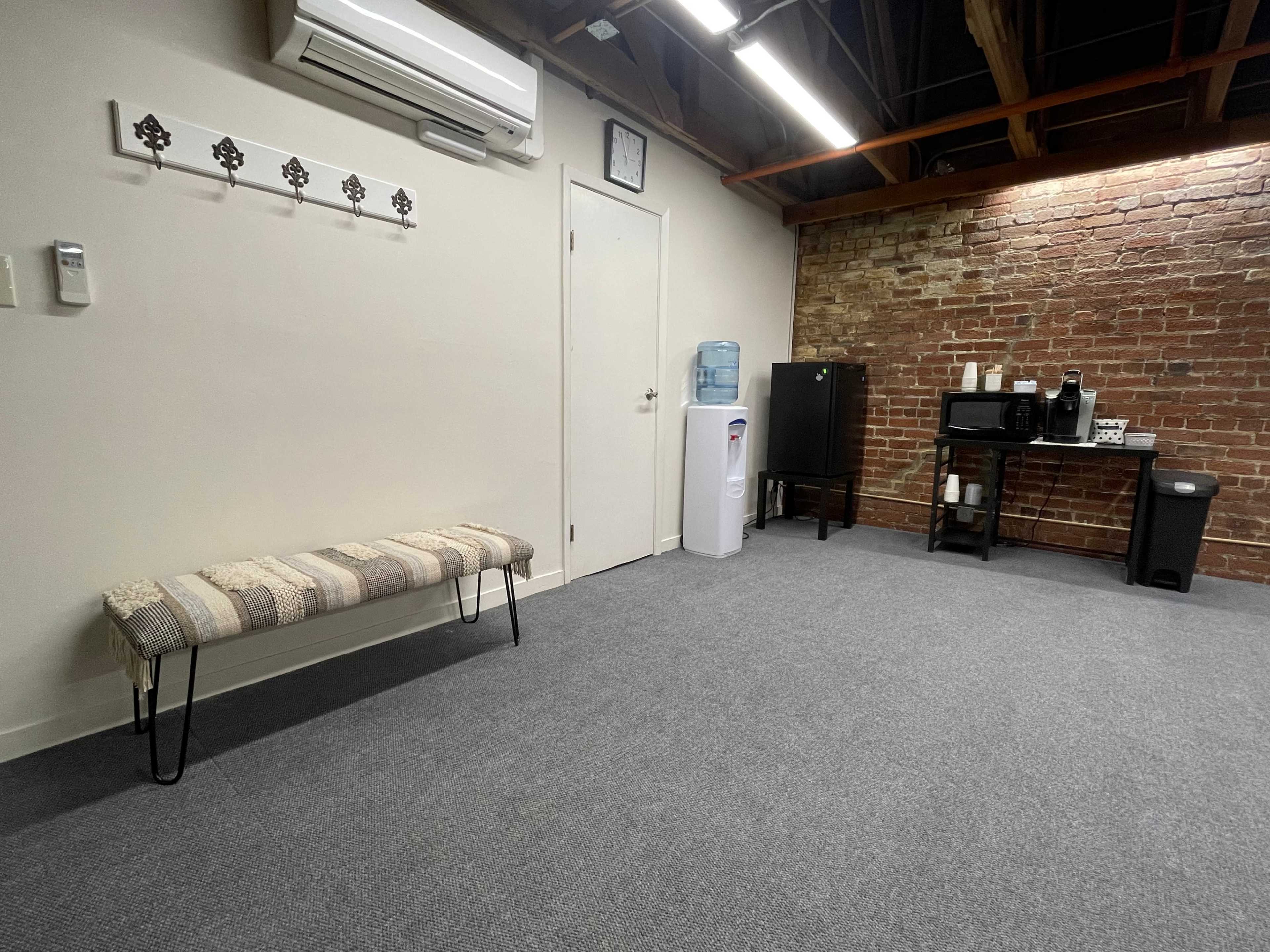 A sparsely furnished room with a bench, a water cooler, a black cabinet, a microwave, and a trash can against a brick wall.