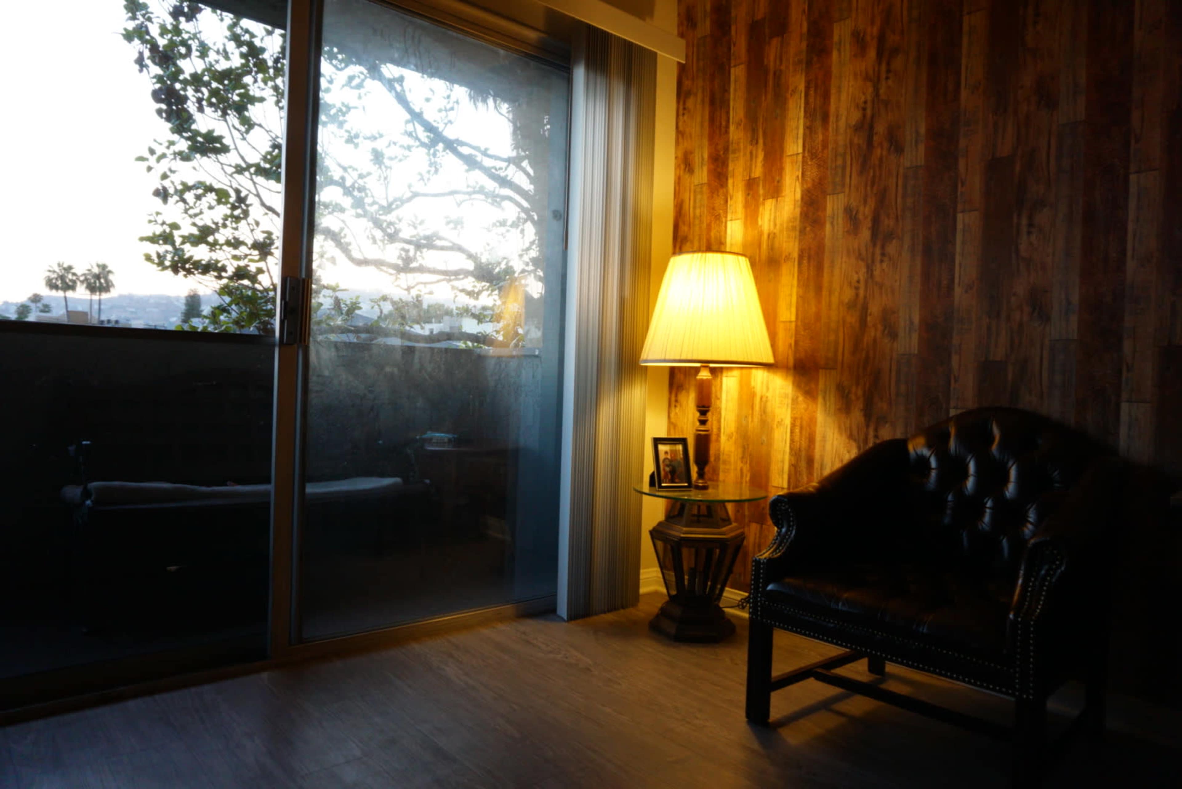 A chair sits beside a lamp on a side table in a room with a wooden wall and large sliding glass door.