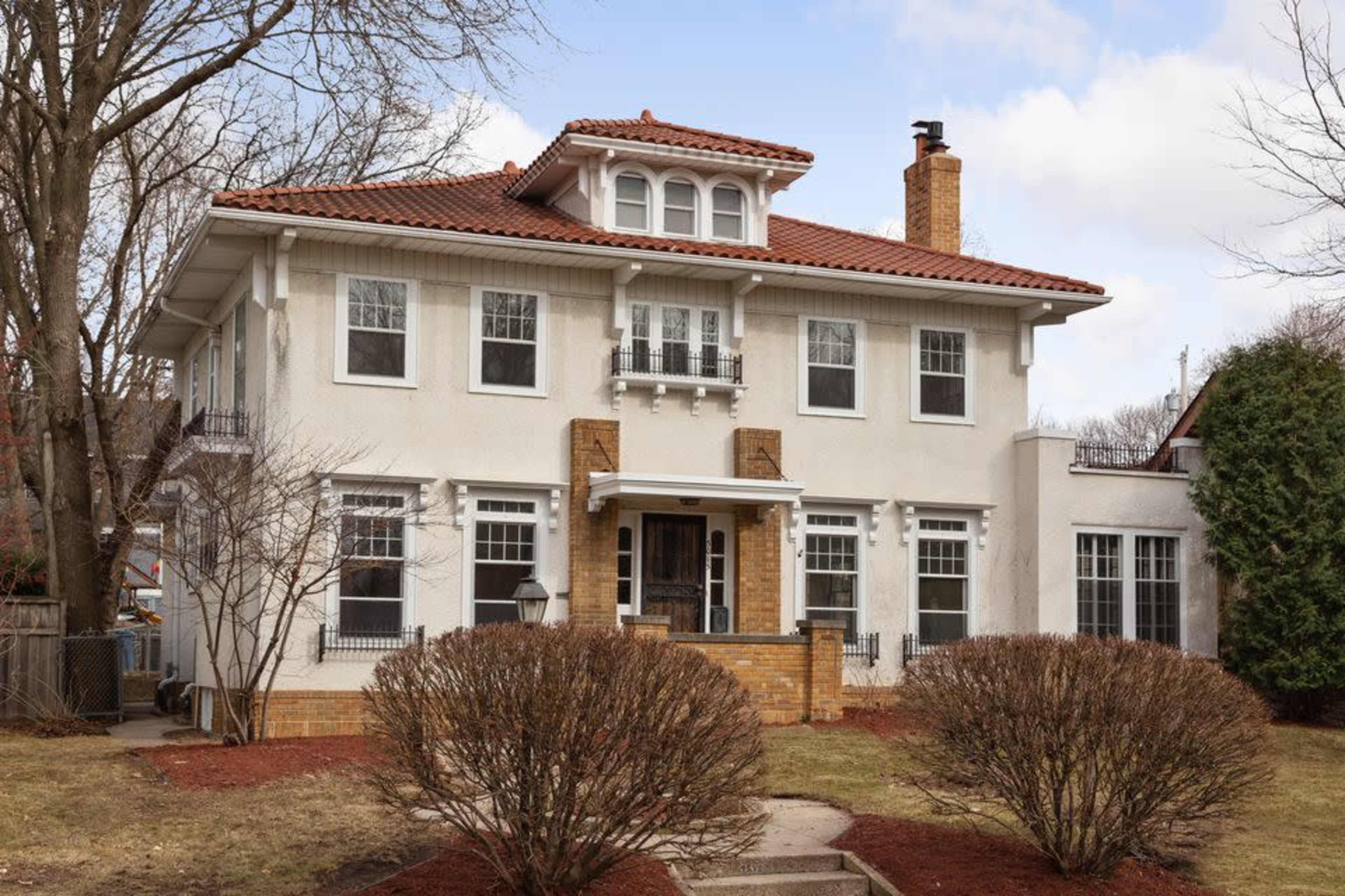 A two-story, cream-colored house with a red tile roof and a brick chimney, surrounded by manicured shrubs and a clear sky.