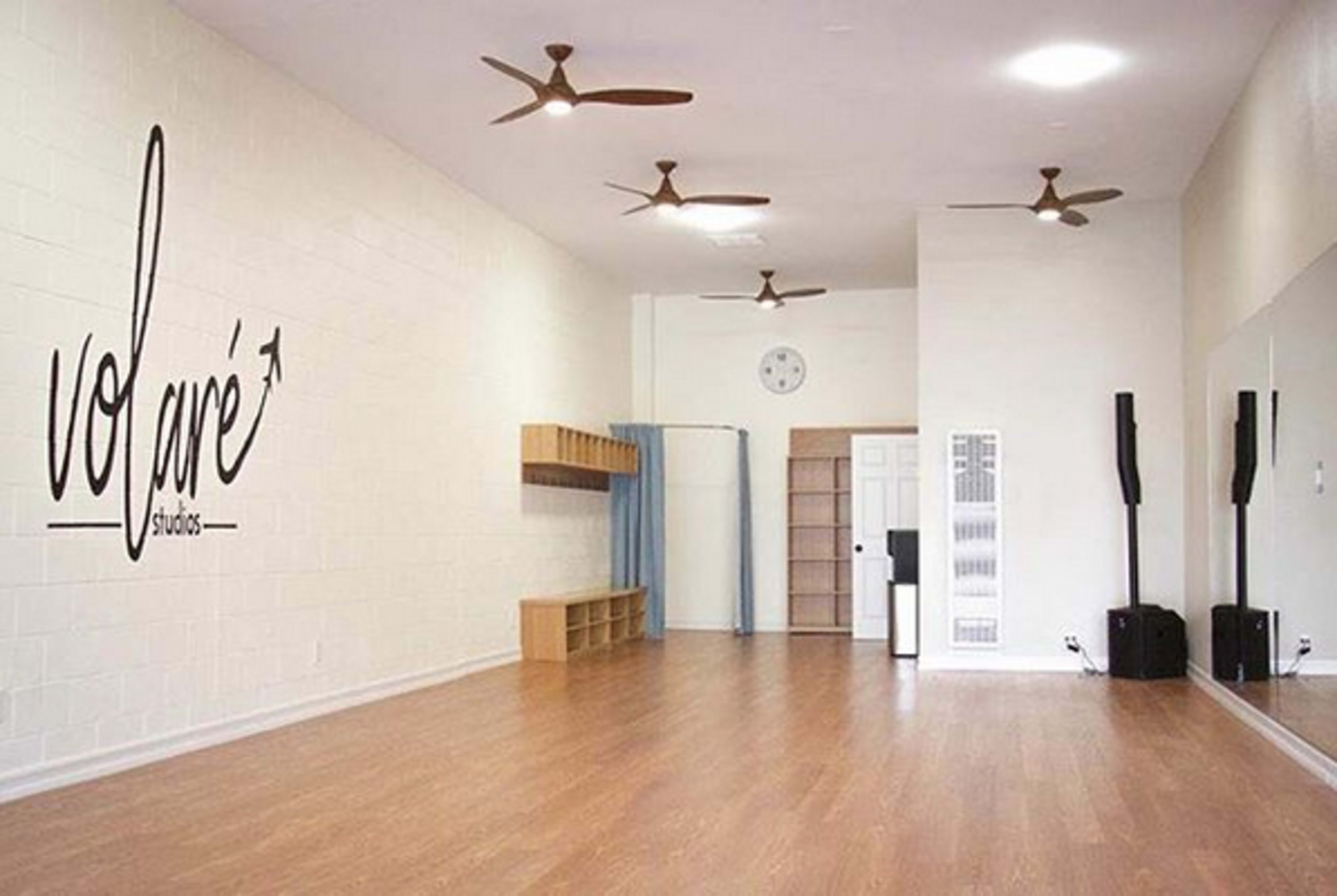 The image shows an empty dance studio with light-colored walls, wooden flooring, and a large wall-mounted mirror.