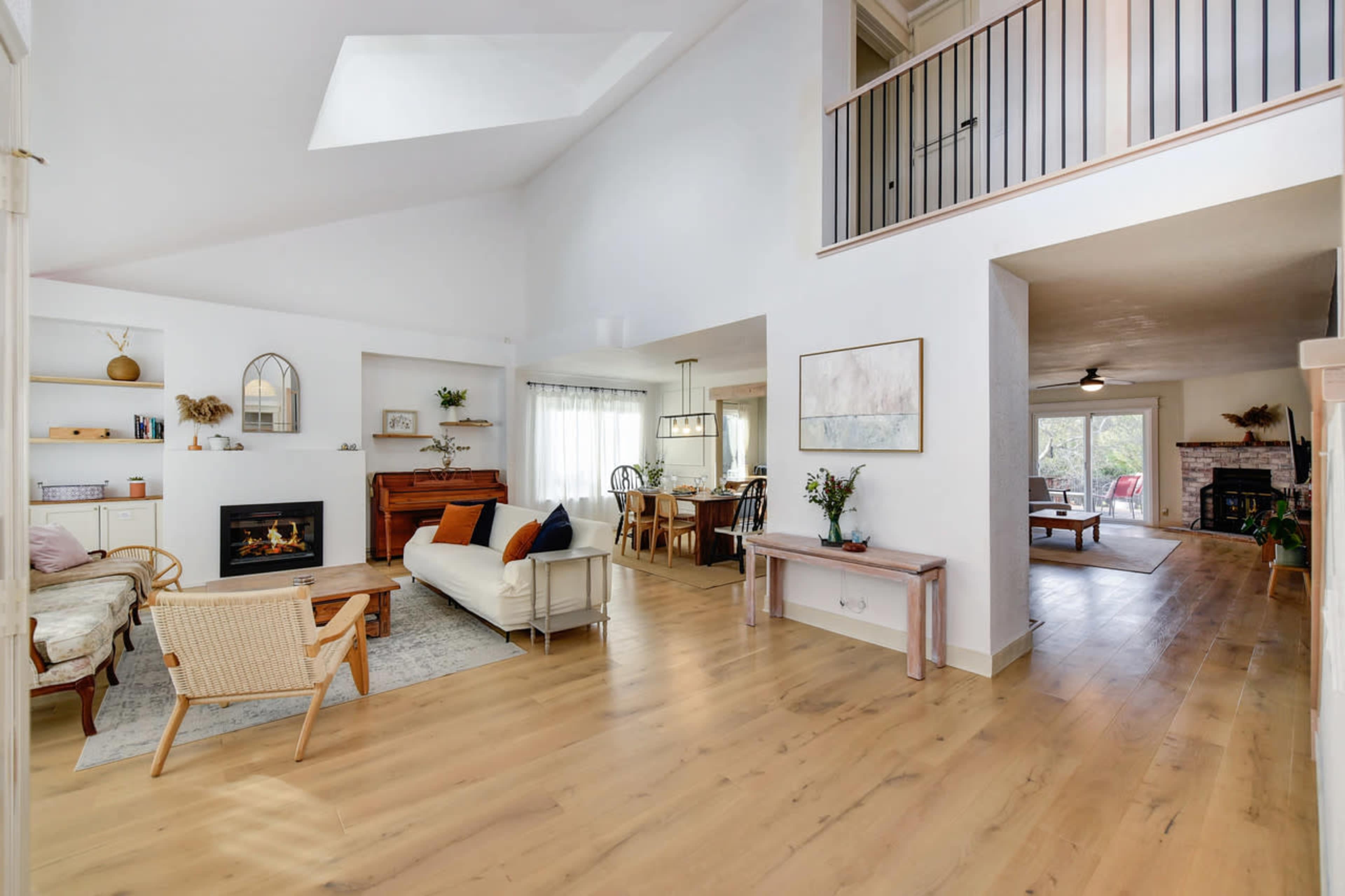 The image shows a spacious, well-lit living area with a white sofa, wooden furniture, and a fireplace, leading into a dining area with large windows.