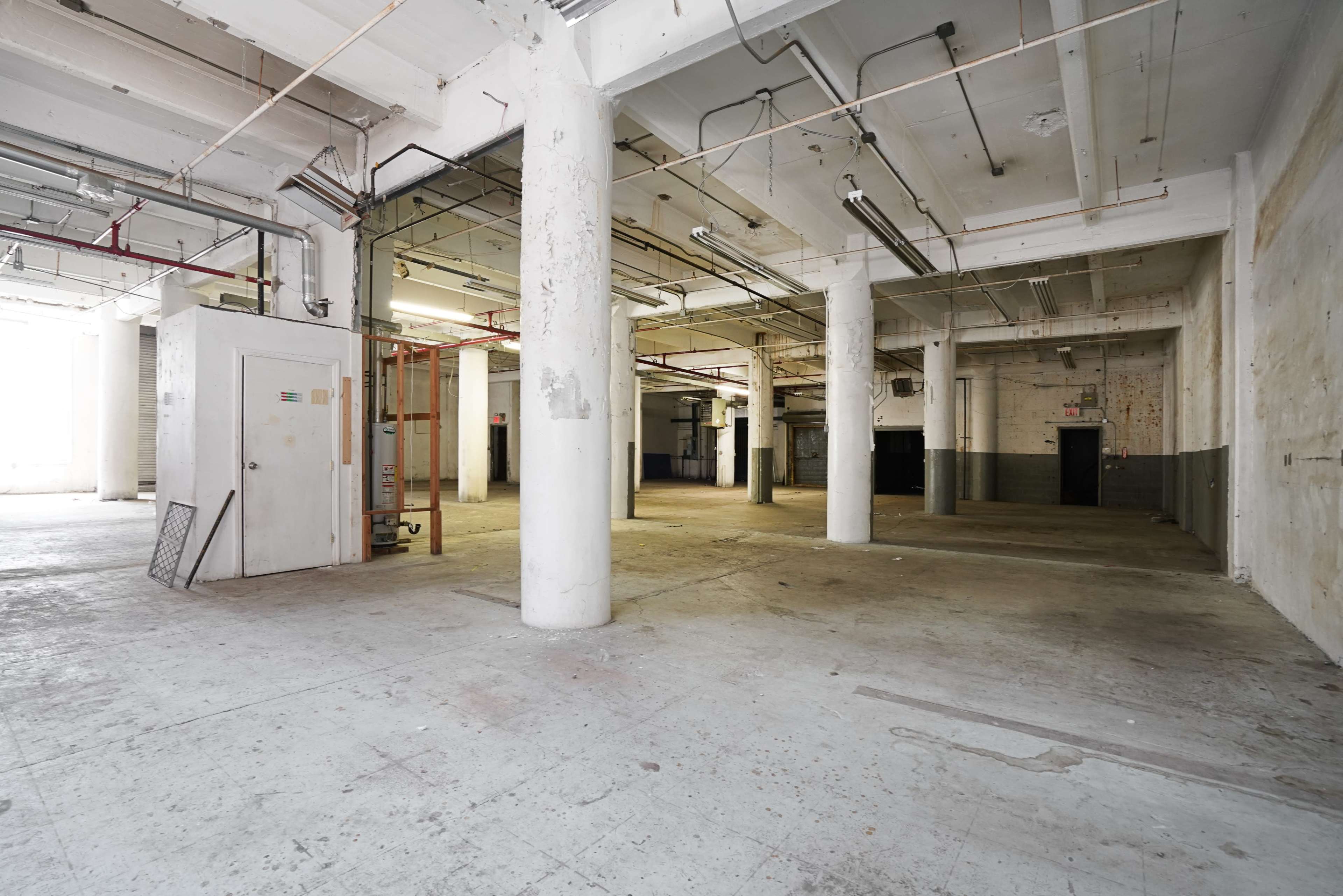 An empty industrial interior with concrete floors, tall columns, and exposed pipes along the ceiling.