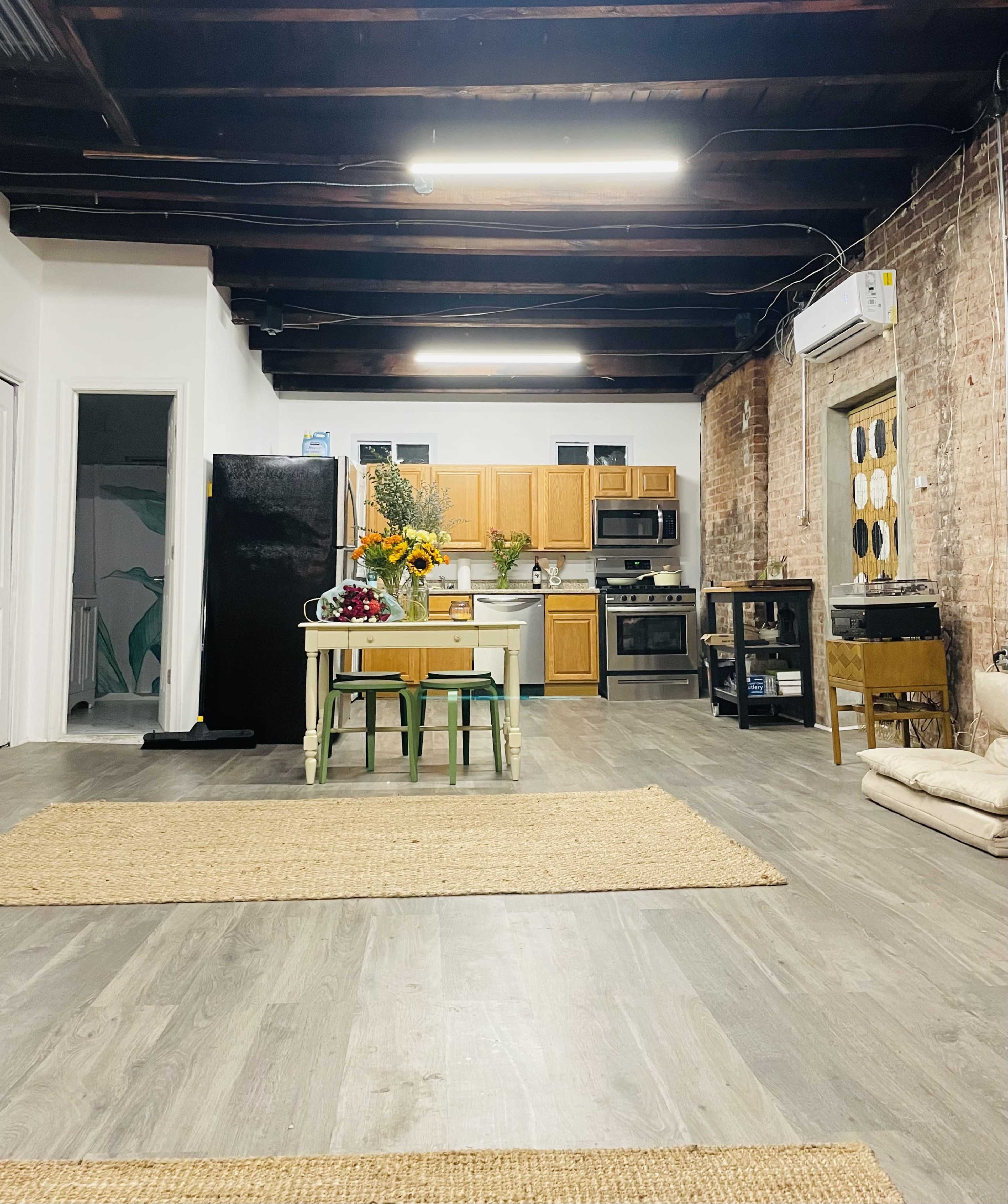 A spacious kitchen area with wooden cabinets, a black refrigerator, and a table set with flowers, all situated in a room with exposed brick walls and wooden beams.