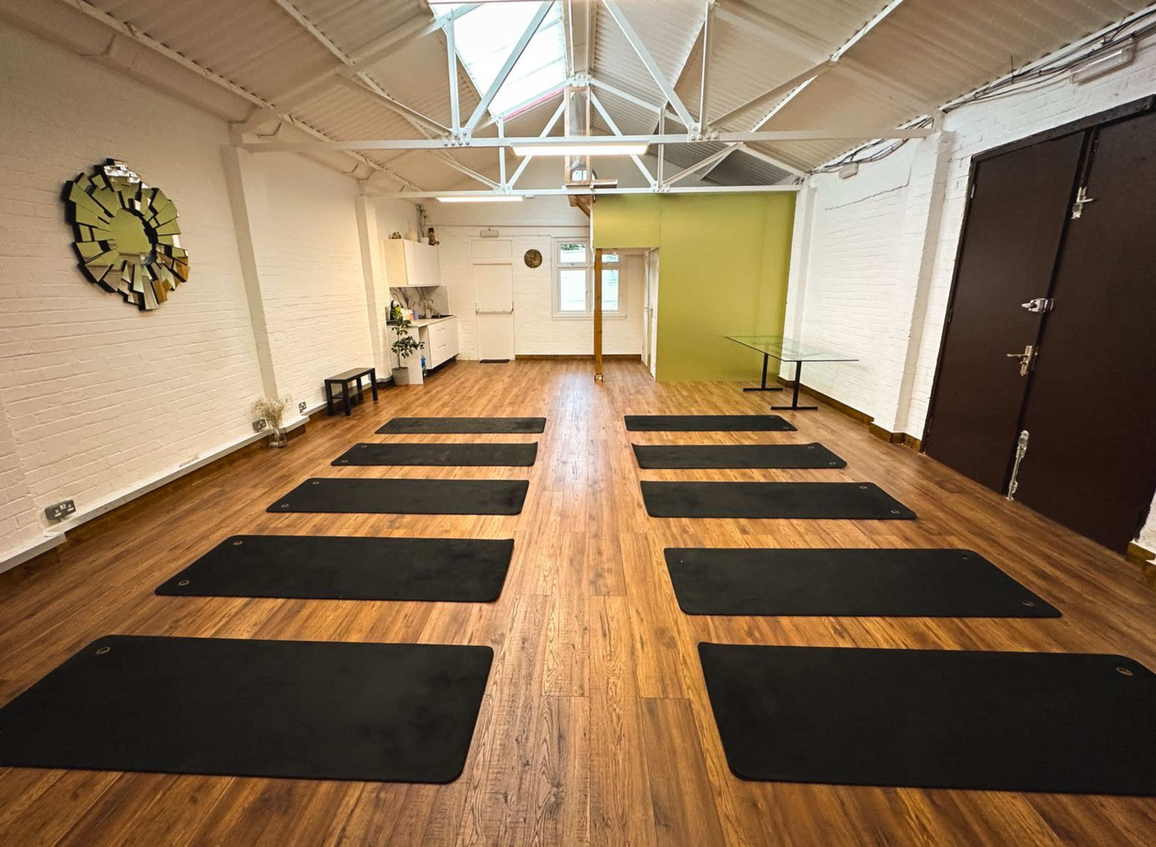 The image shows a spacious studio with wooden floors, featuring several yoga mats arranged in parallel lines beneath a skylighted ceiling.