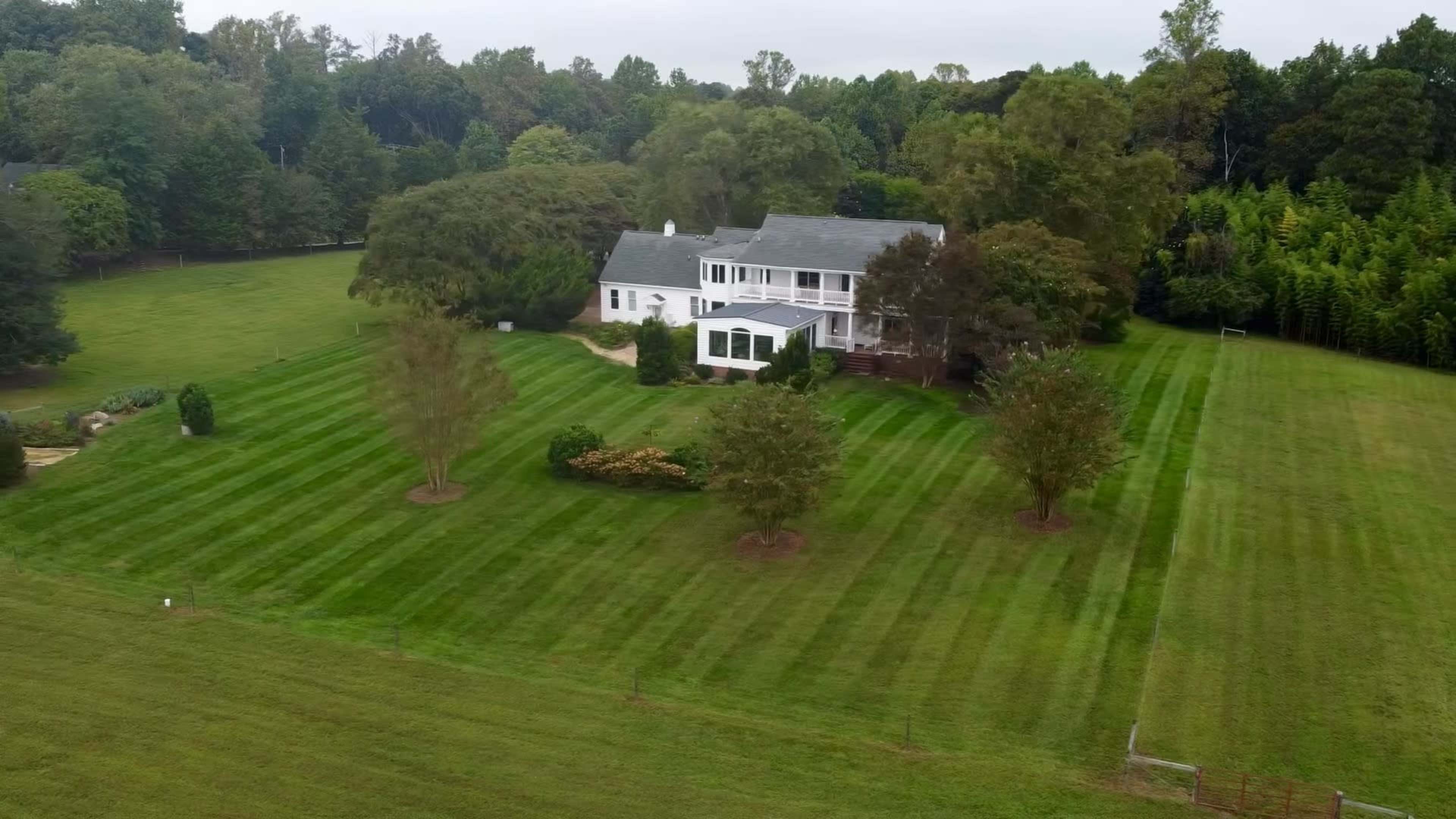 A large, two-story white house is surrounded by neatly striped green lawn and trees on a spacious property.