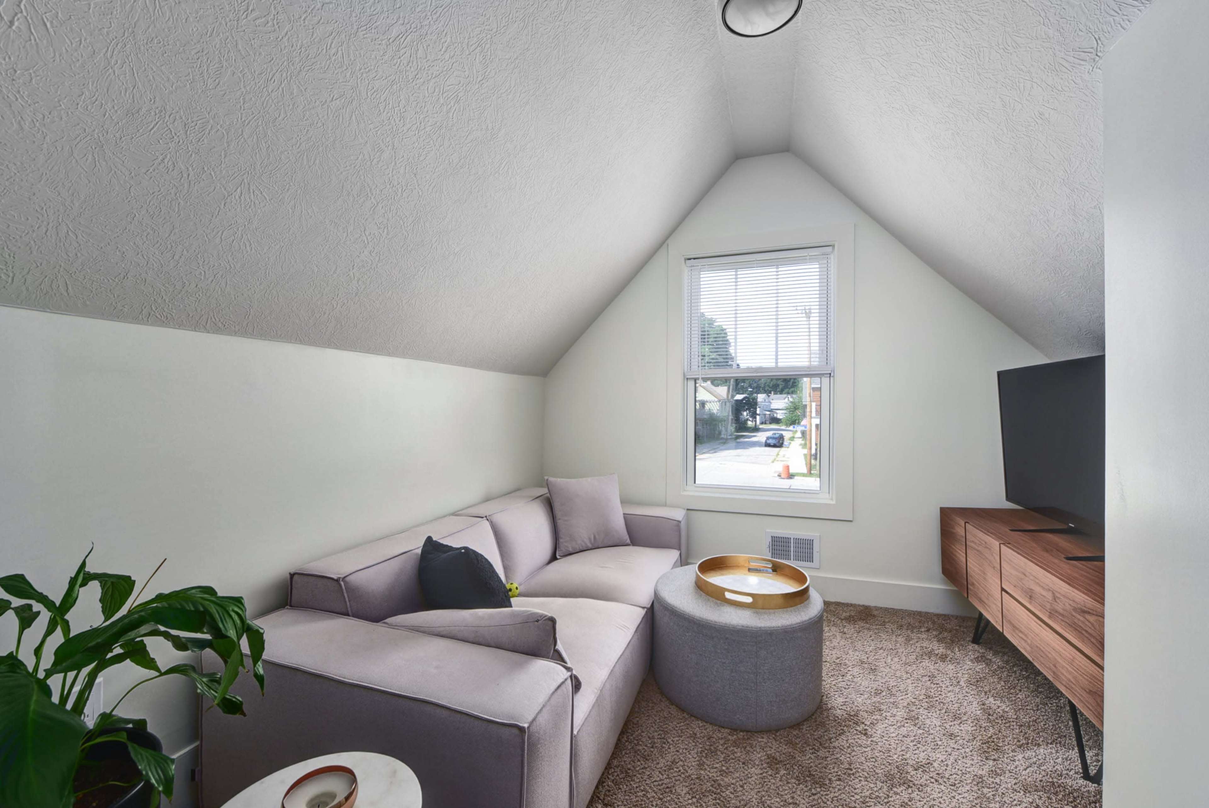 A small living room features a gray sectional sofa, a wooden TV stand, and a round coffee table, with a window allowing natural light to enter.
