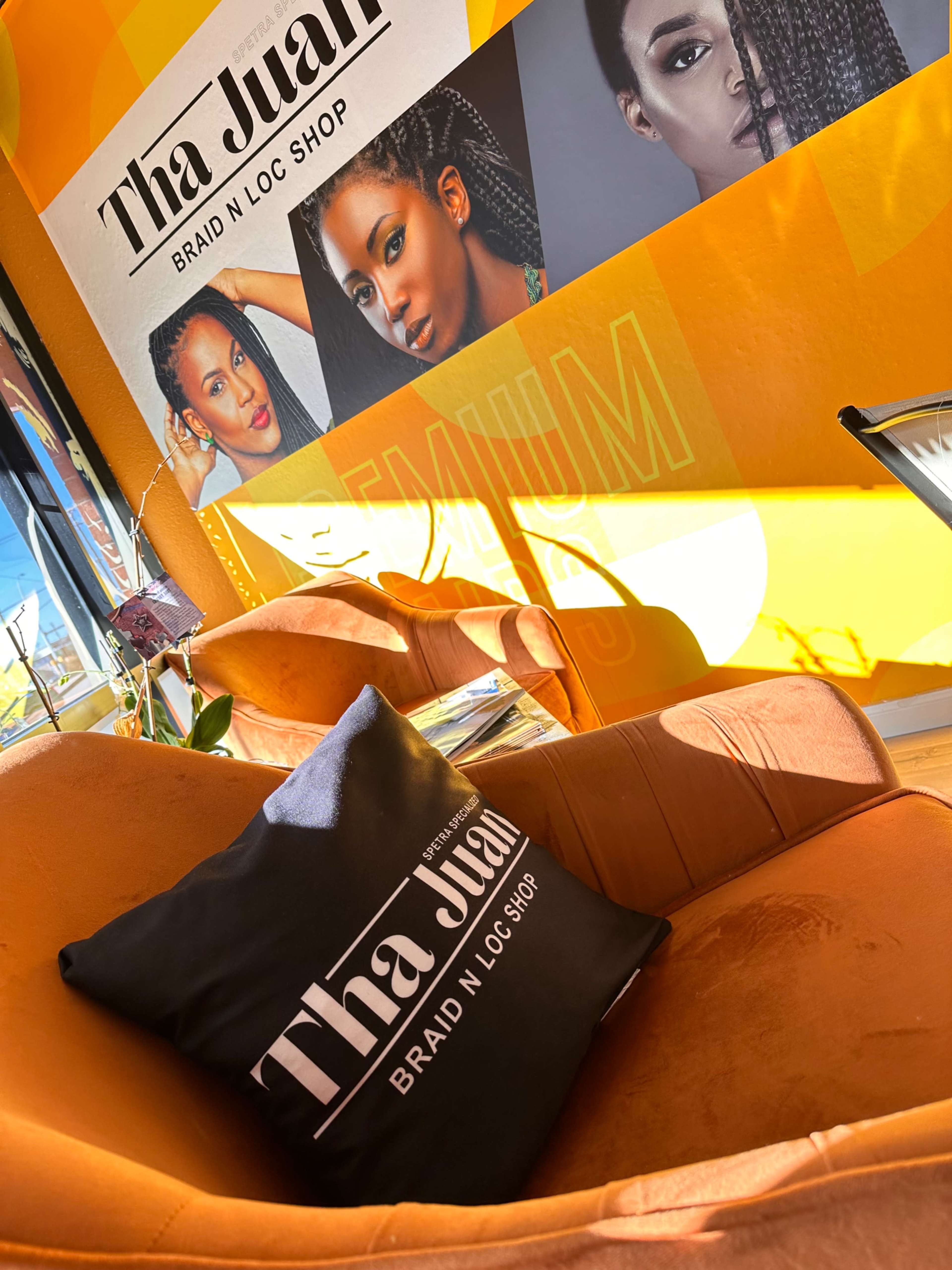 A chair with a black cushion labeled "Tha Juan Braid N Loc Shop" is placed against a bright orange wall featuring images of individuals with braided hairstyles.
