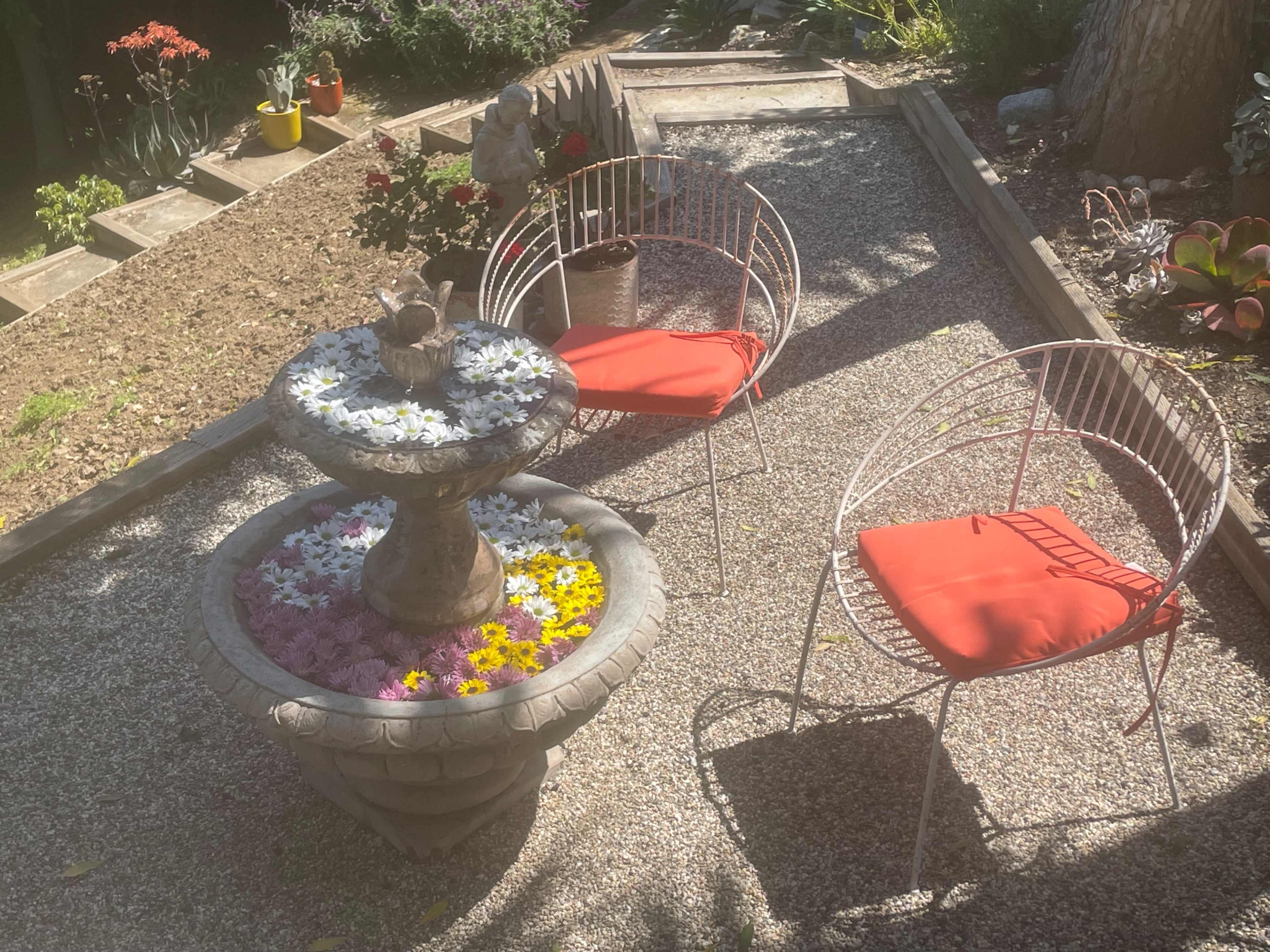 A garden scene features a stone fountain surrounded by colorful flowers and two white chairs with orange cushions.