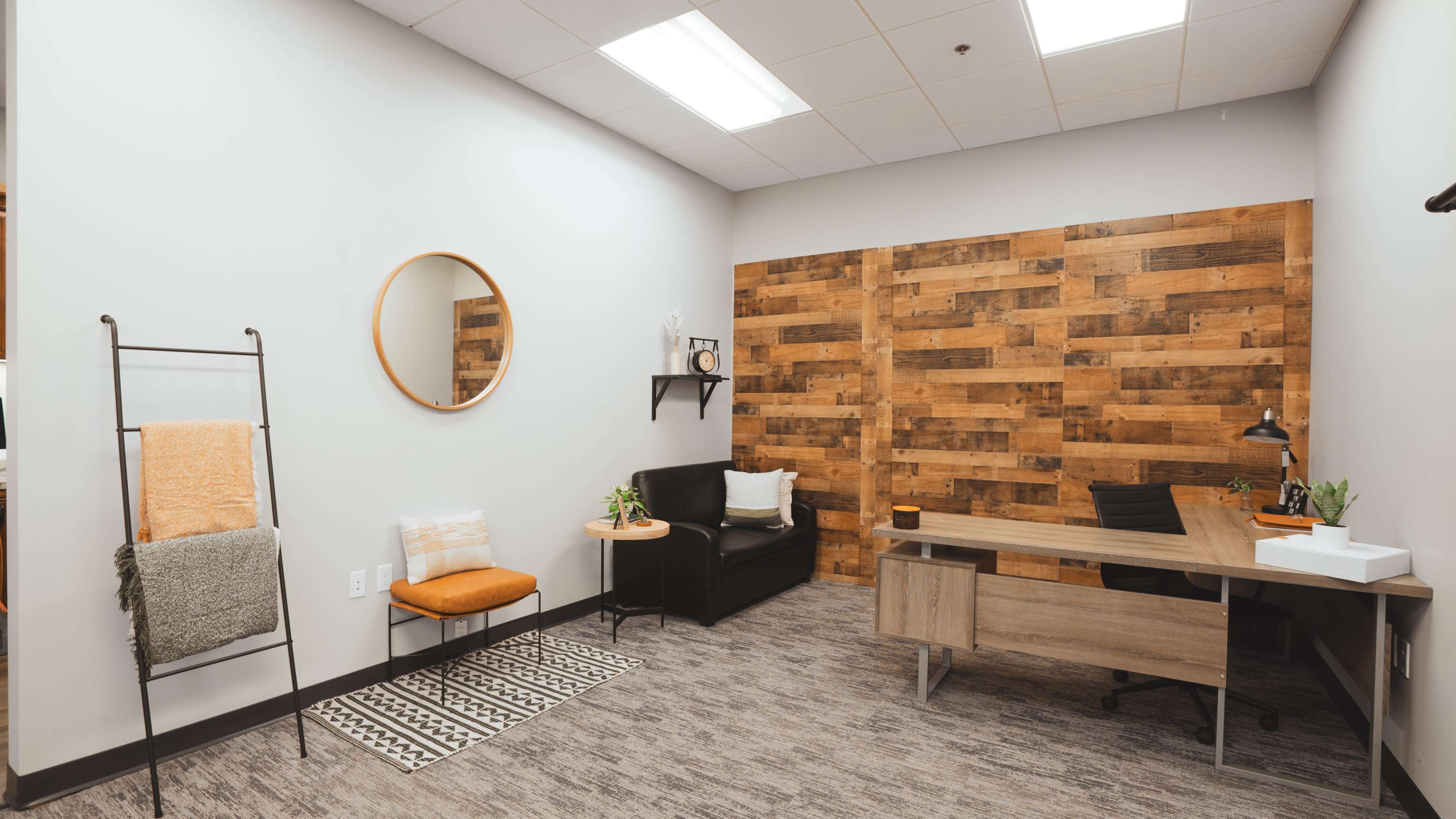 A modern office space featuring a wooden accent wall, a desk with a chair, a black sofa, a small coffee table, and a decorative mirror.