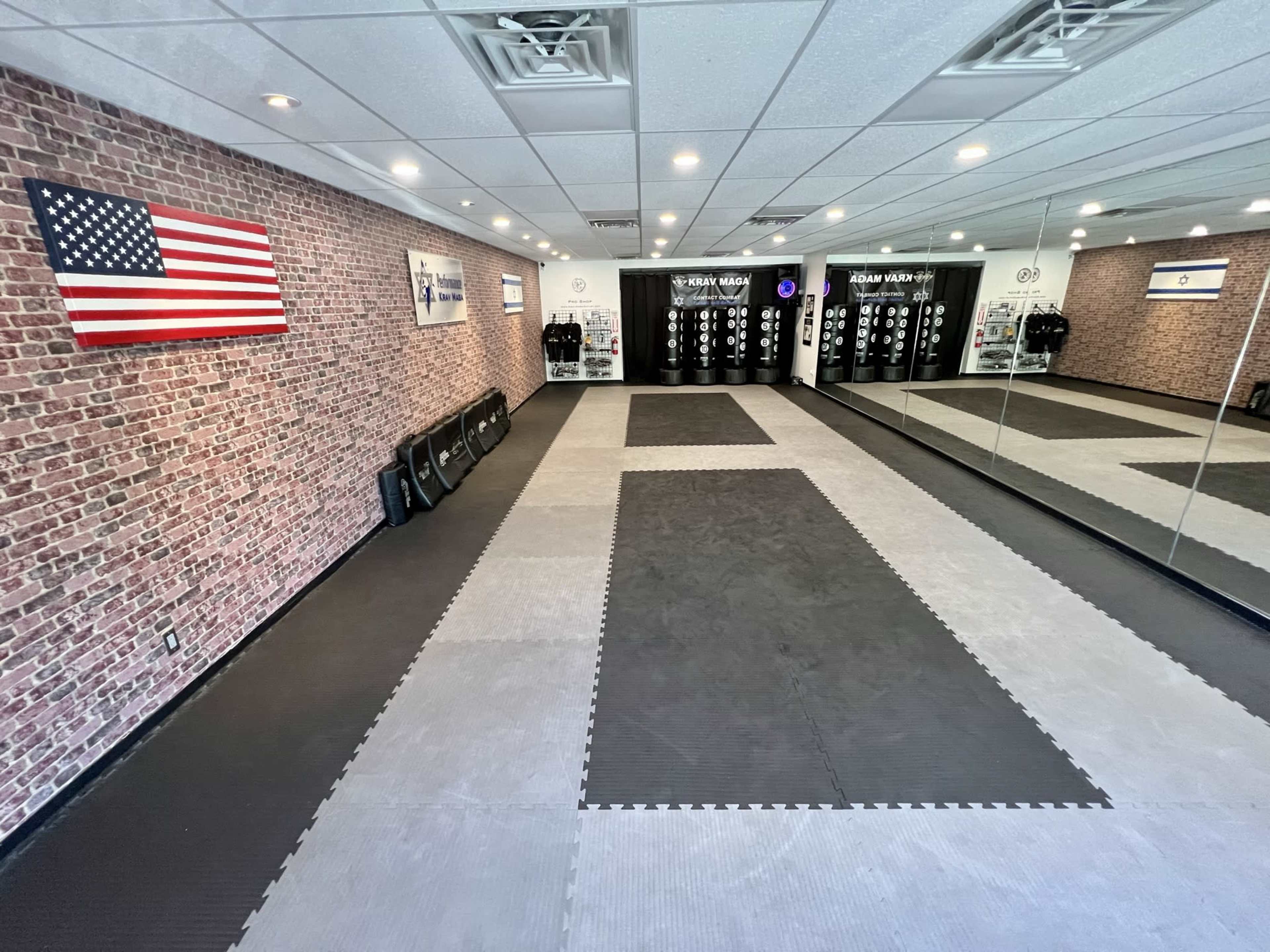 A spacious martial arts studio with a brick wall, an American flag, and mats on the floor, along with various training equipment displayed.