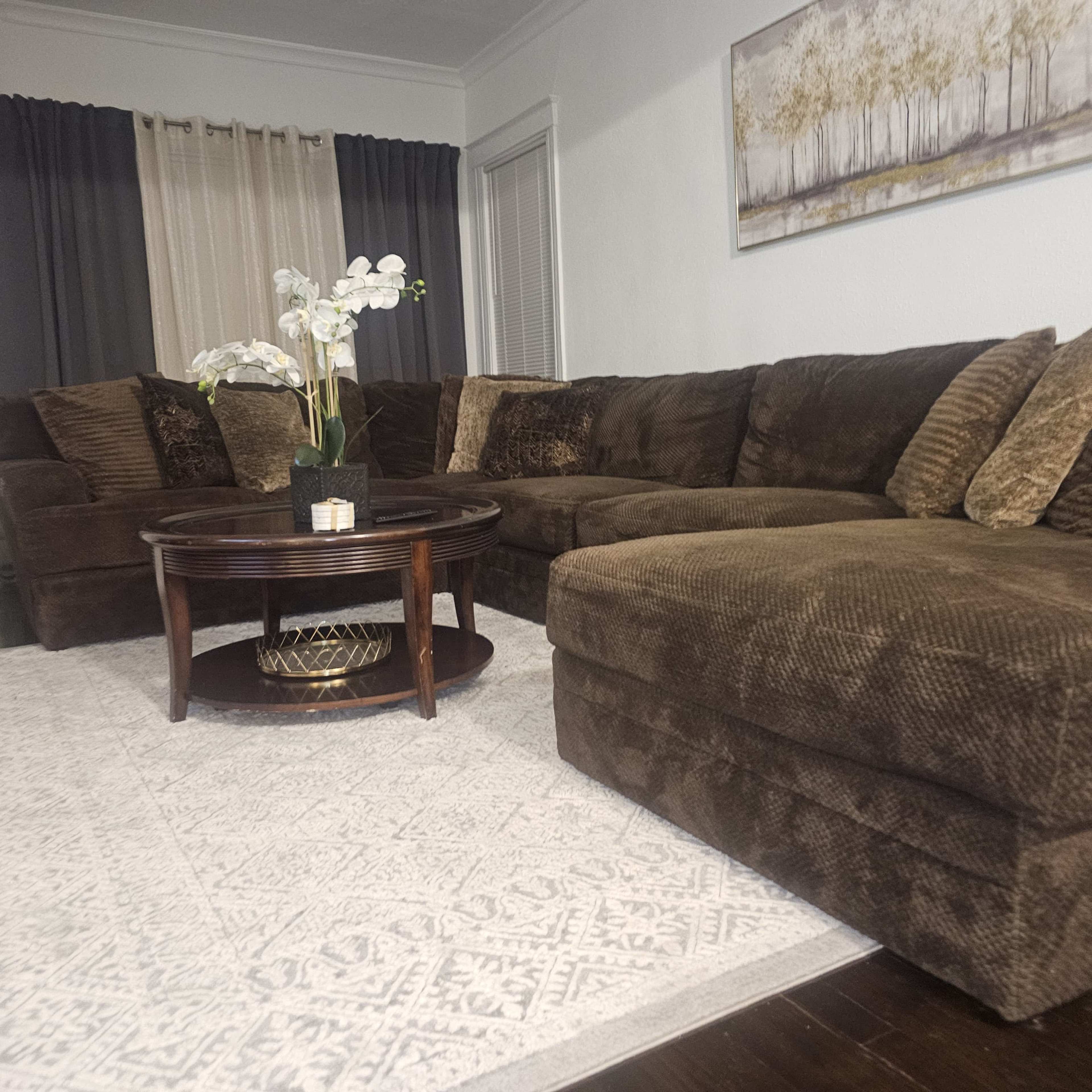 The image shows a living room with a brown sectional sofa, a round wooden coffee table, and a decorative plant on the table, set against a light-colored wall with curtains.