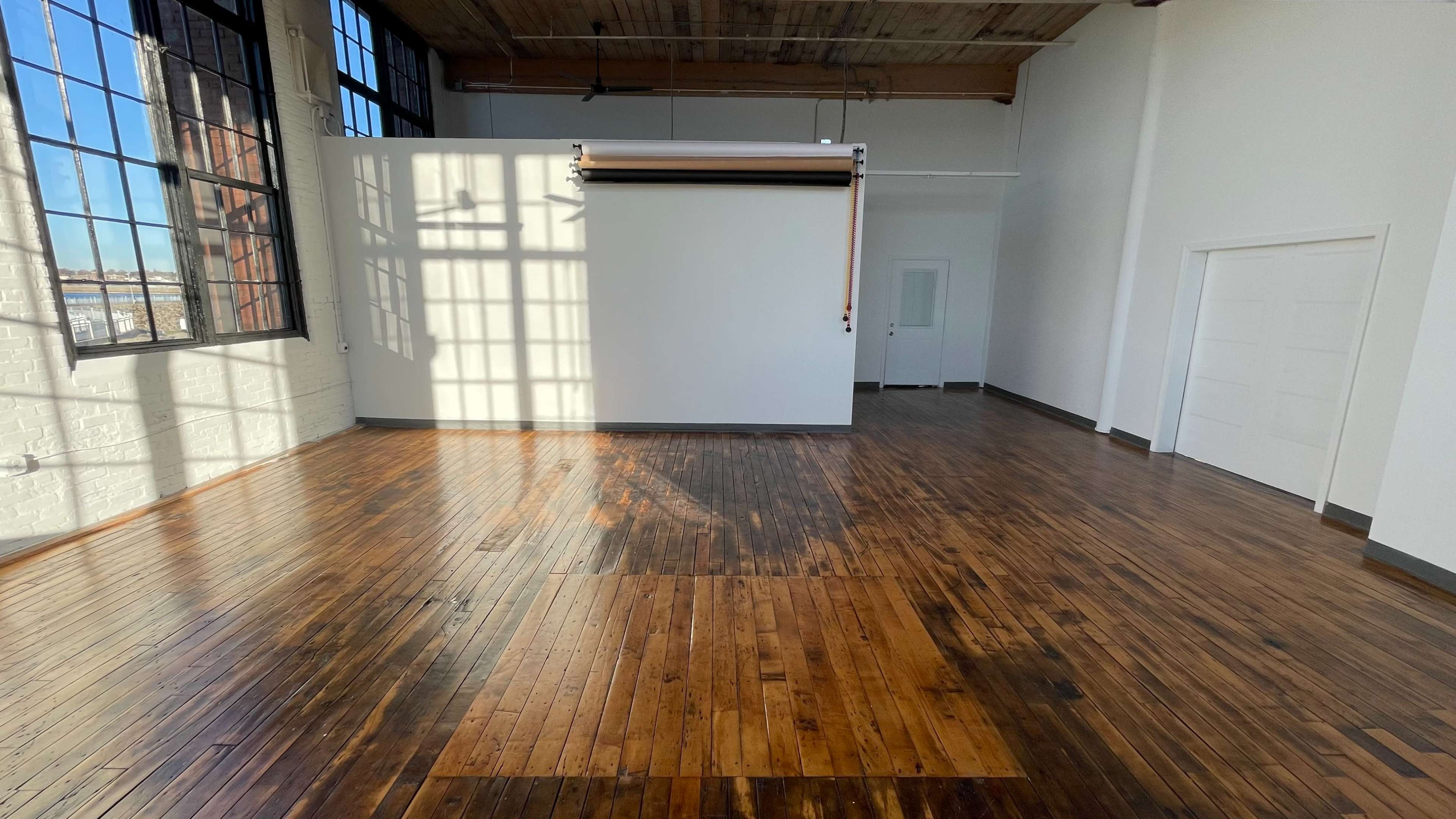 A spacious, empty room with large windows, hardwood floors, and a white wall featuring a roll-up backdrop.