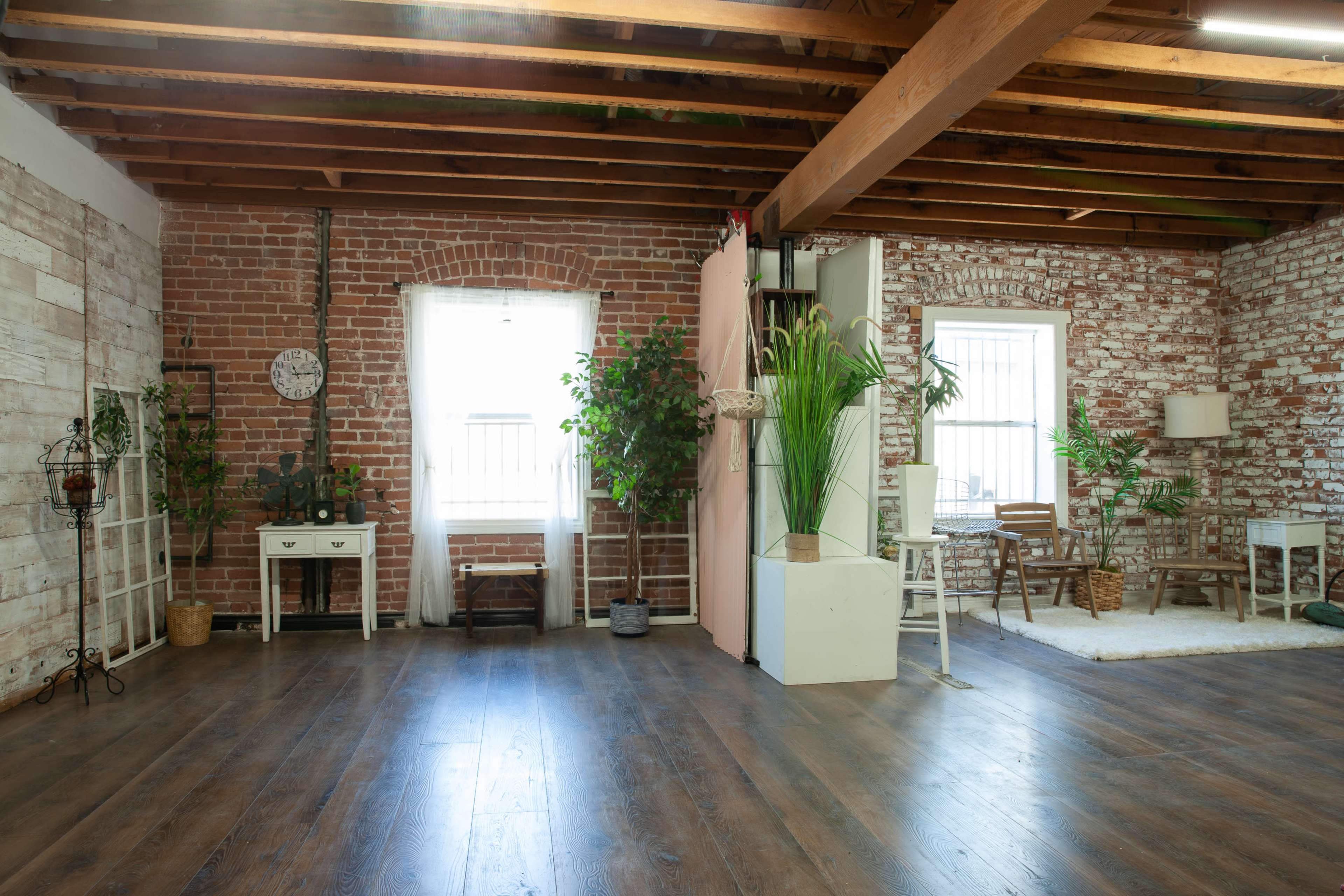 A spacious, well-lit interior with exposed brick walls, wooden beams, and various plants, along with furniture arranged in a cozy setting.