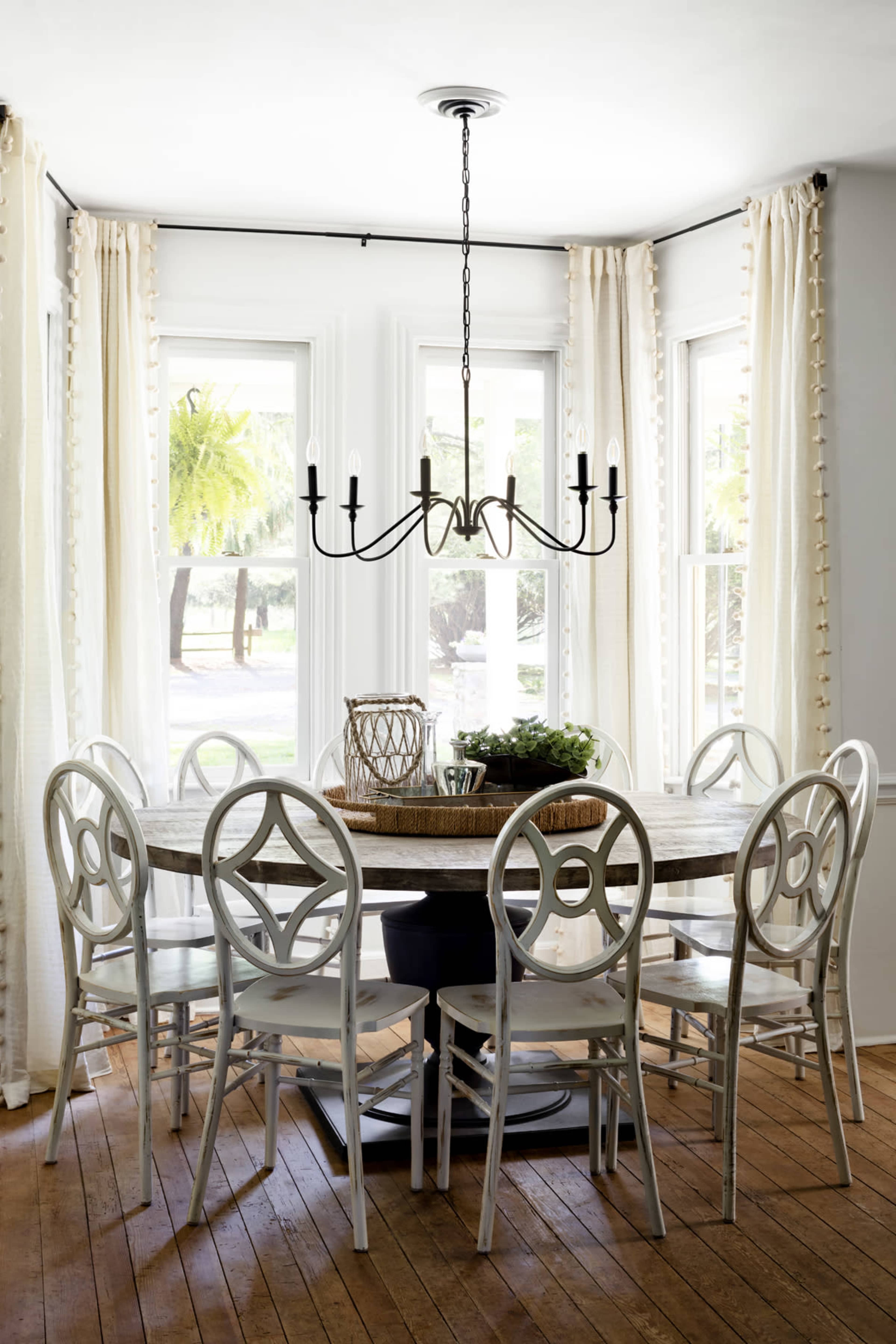A round dining table surrounded by eight chairs is set in a bright room with large windows adorned with sheer curtains.