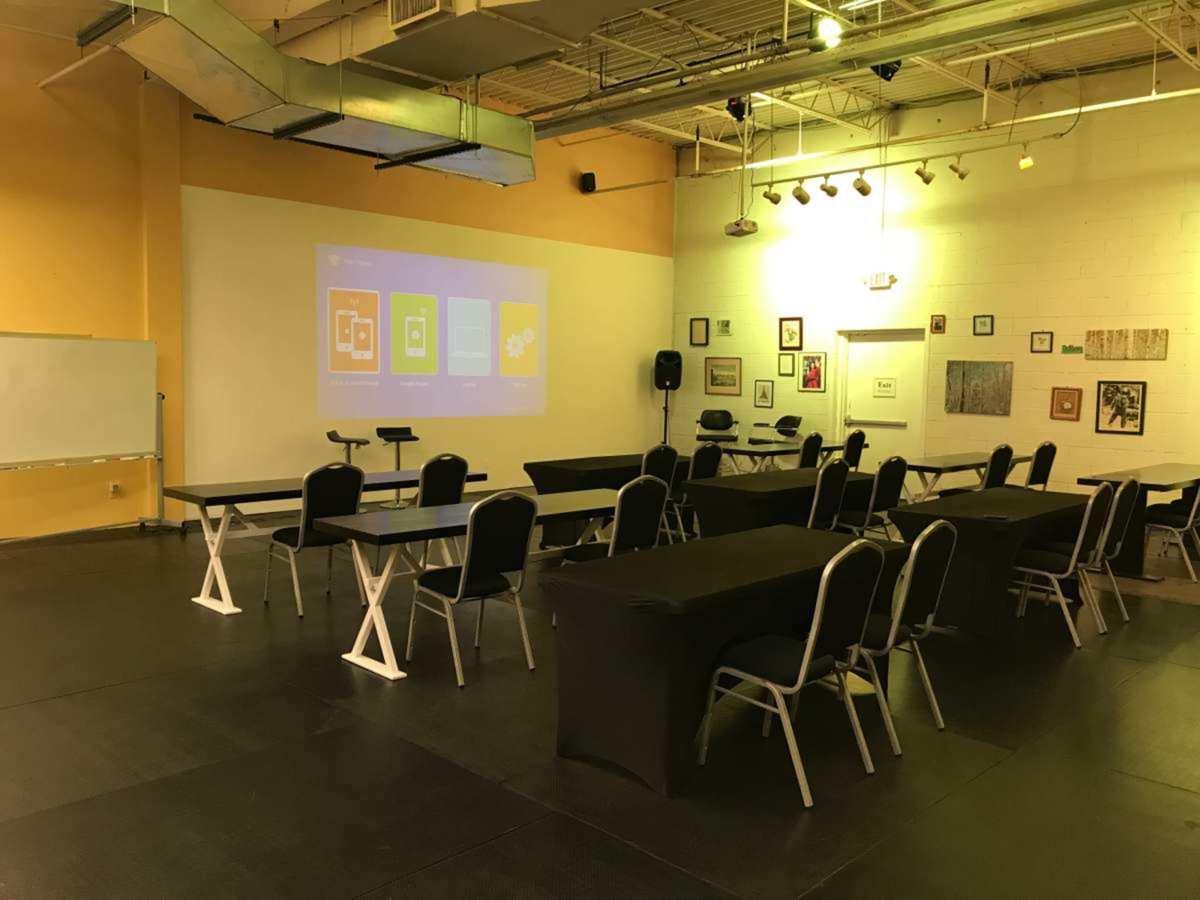 The image shows a conference room setup with several tables and chairs arranged in a U-shape, facing a projection screen on a yellow wall.